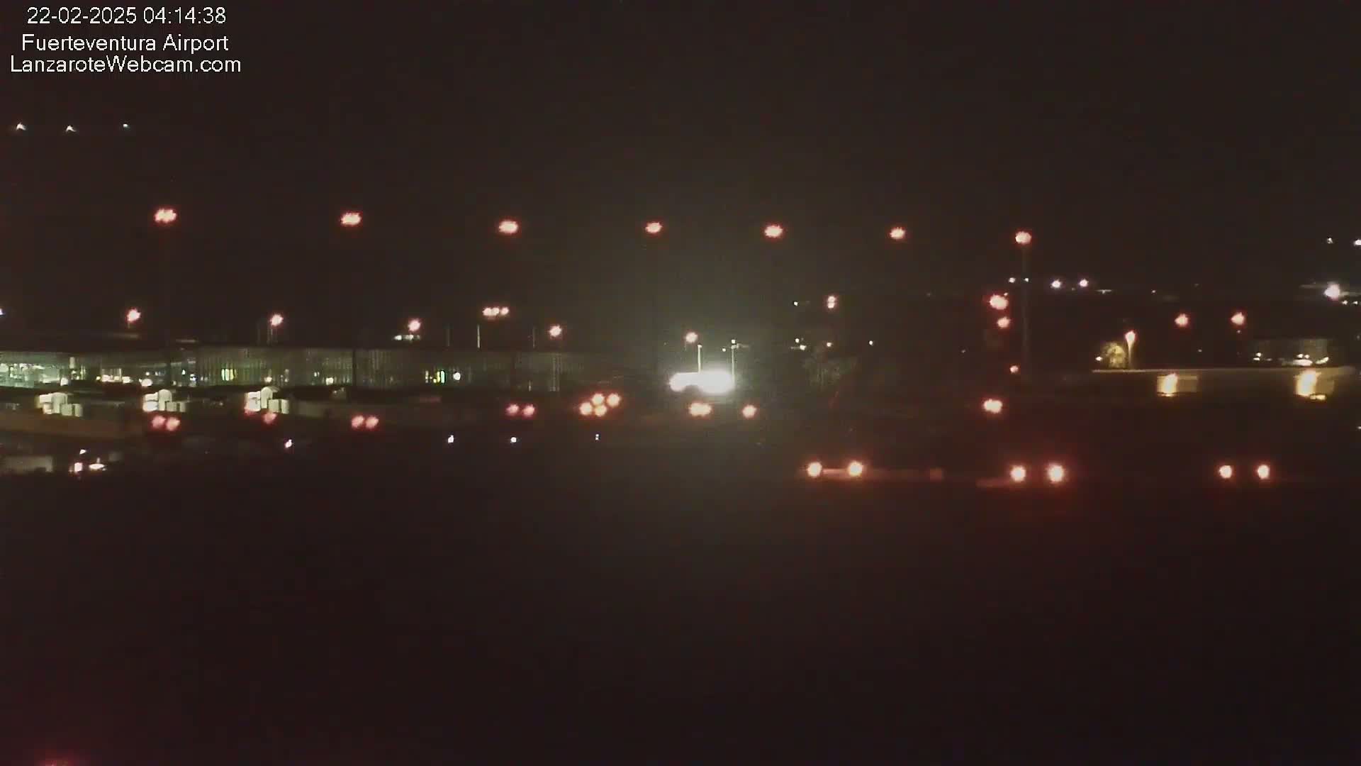 A nighttime long shot shows an airport runway with numerous lights illuminating the tarmac and surrounding buildings under a clear, dark sky.