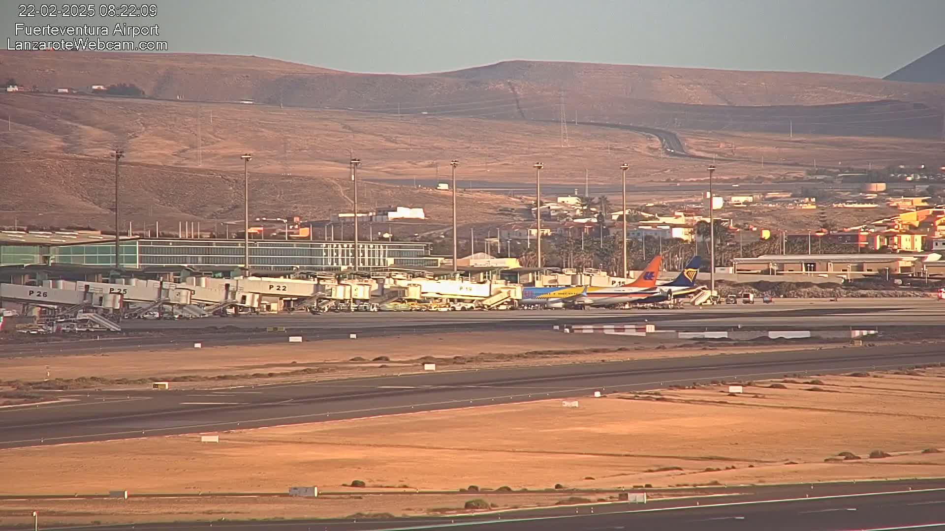 Several airplanes are parked at gates of an airport terminal under a clear, light-orange sky, with brown hills visible in the background.