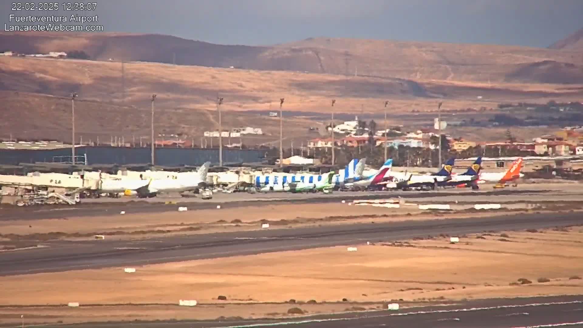 Several airplanes are parked at an airport tarmac under a mostly clear sky, with a dry, brown landscape visible in the background.