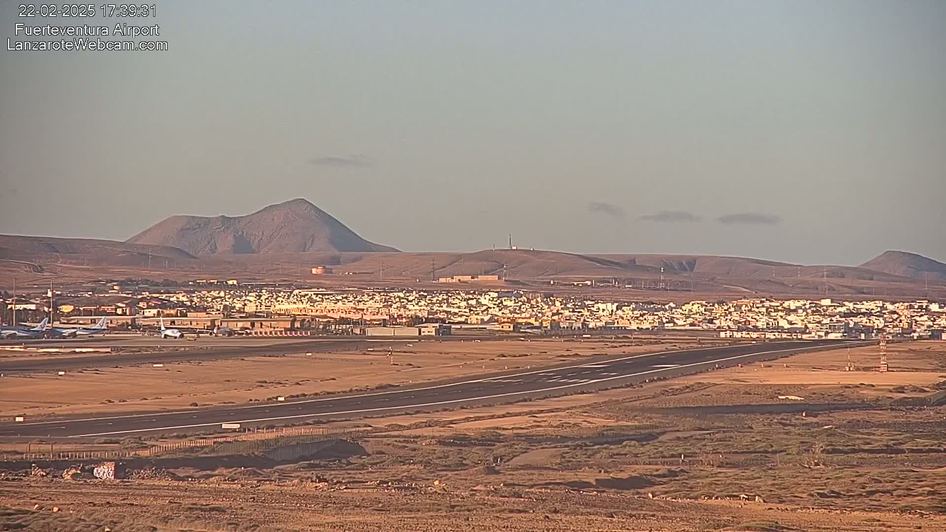 A long runway at an airport is shown with several planes on the tarmac, a town in the background, and distant volcanic hills under a clear, light-colored sky.