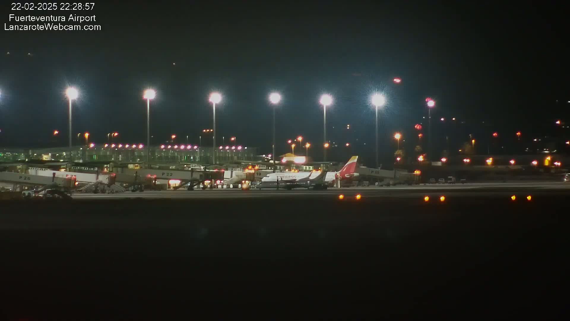 At night, several airplanes are parked at gates in an airport, illuminated by bright lights.