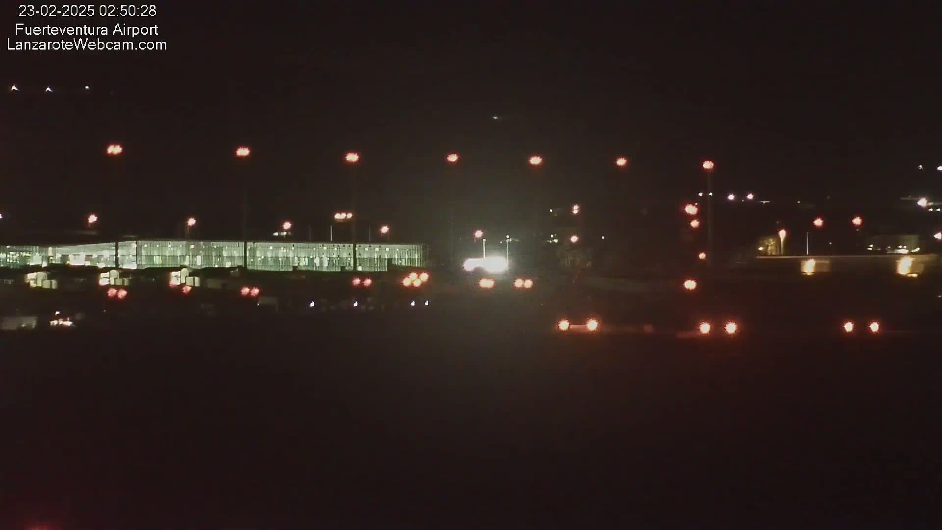A nighttime long shot of a brightly lit airport terminal and roadway with vehicles, under a clear, dark sky.