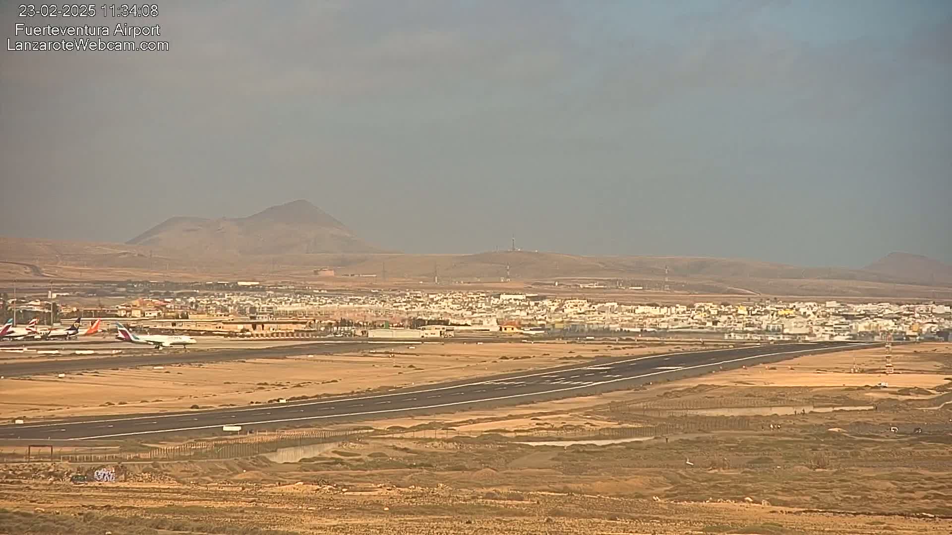 A long airport runway stretches across a flat, arid landscape under an overcast sky, with several airplanes parked near the terminal and a town visible in the distance.