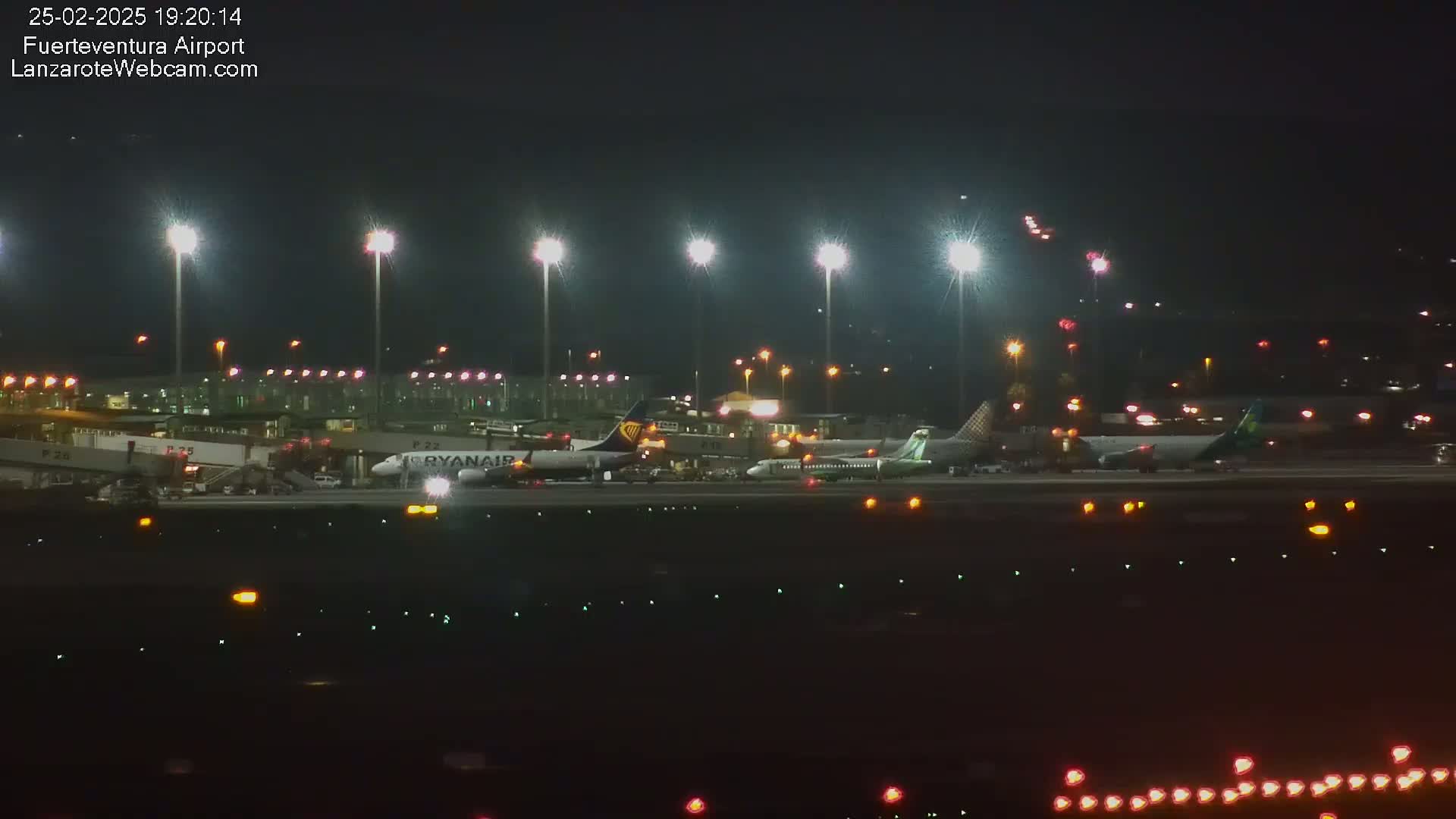 At night, several airplanes are parked at a brightly lit airport with numerous lights illuminating the tarmac.