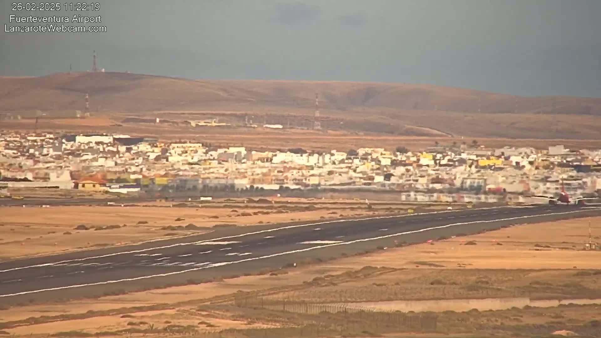 An airport runway stretches across a flat, arid landscape toward a distant town under an overcast sky.