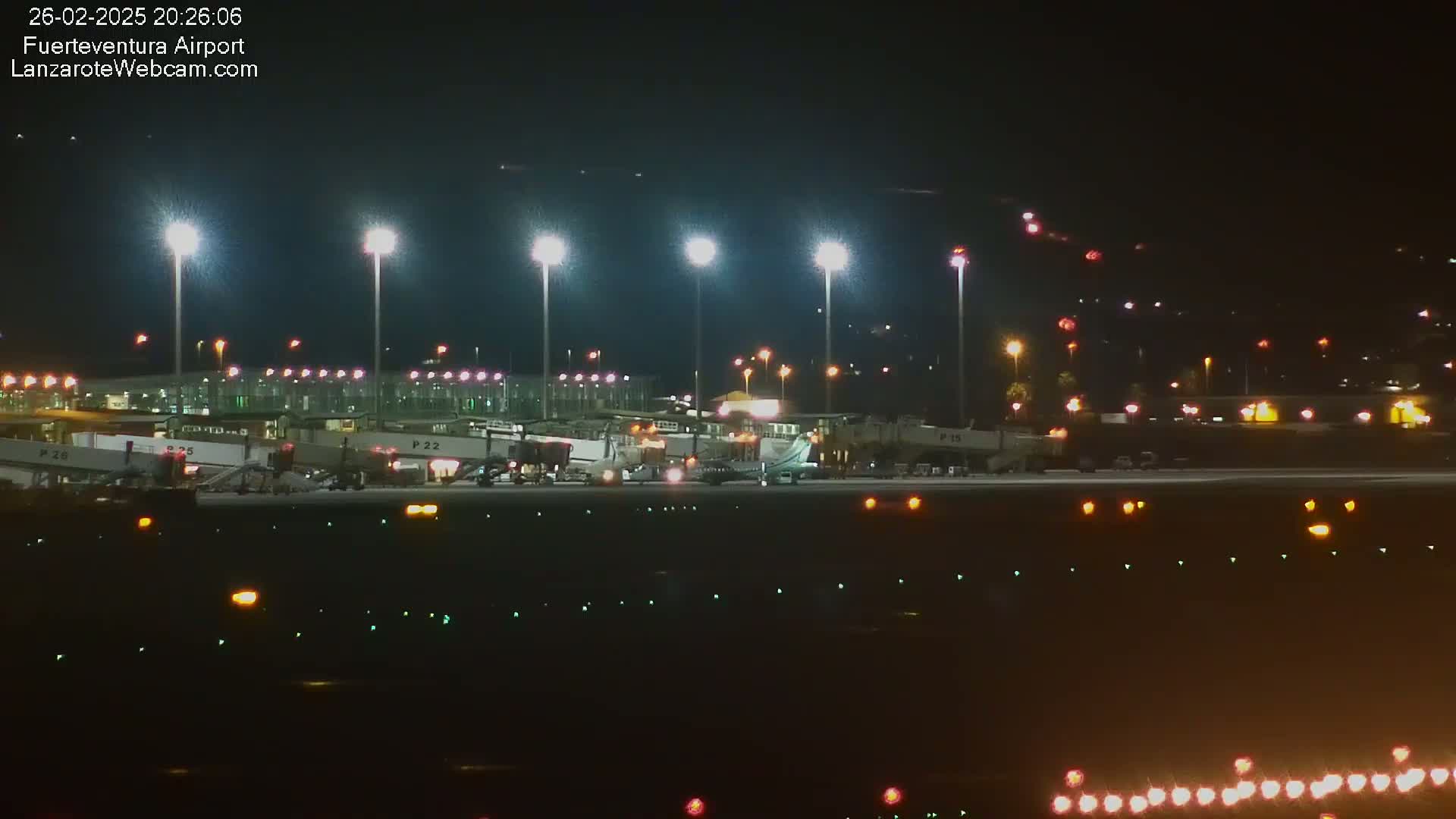 At night, a brightly lit airport tarmac shows several airplanes parked at gates with ground support vehicles nearby.