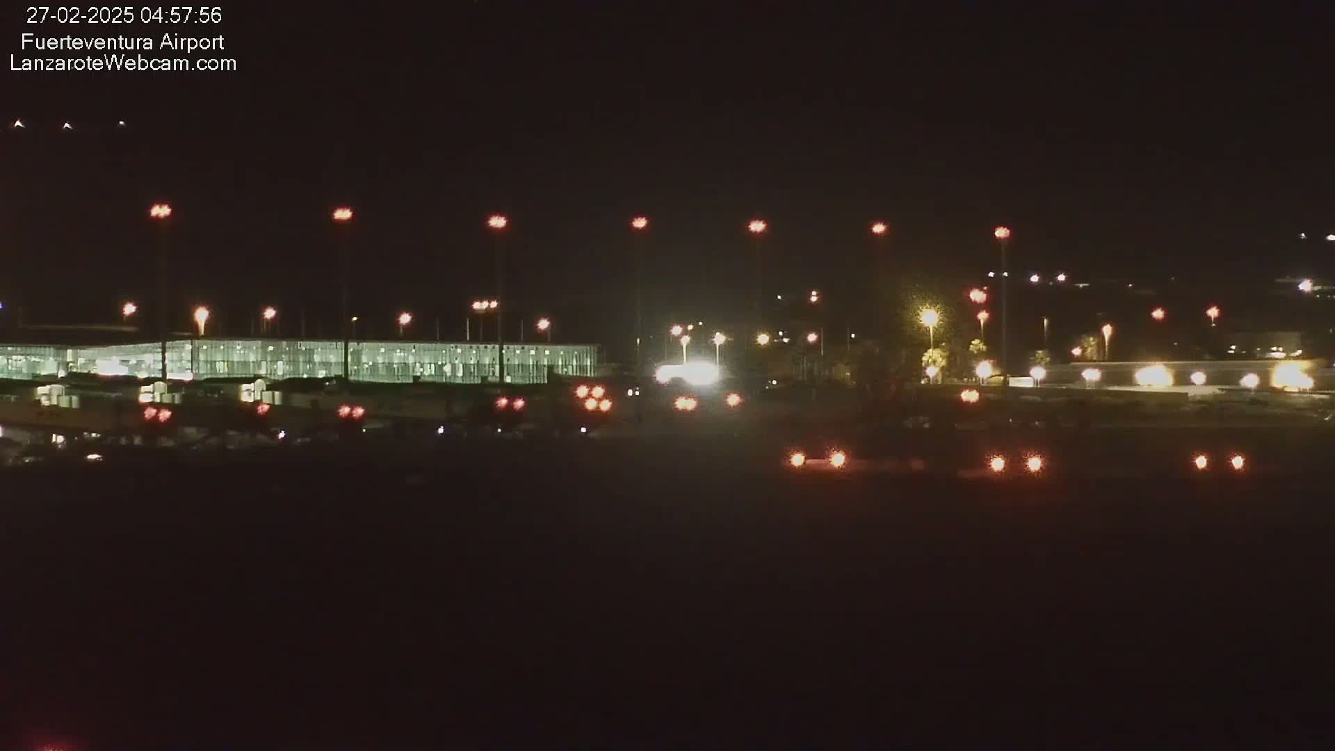 A brightly lit airport terminal at night, with vehicles visible in the foreground and faint lights in the background under a clear, dark sky.