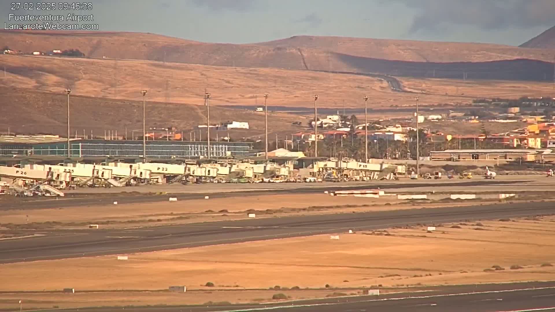 An airport tarmac with several jet bridges connected to a terminal building, under a mostly clear sky, with arid hills in the background.