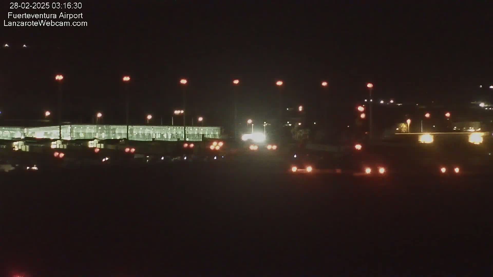 A nighttime long shot of an airport terminal with vehicles and lights visible under a dark sky.