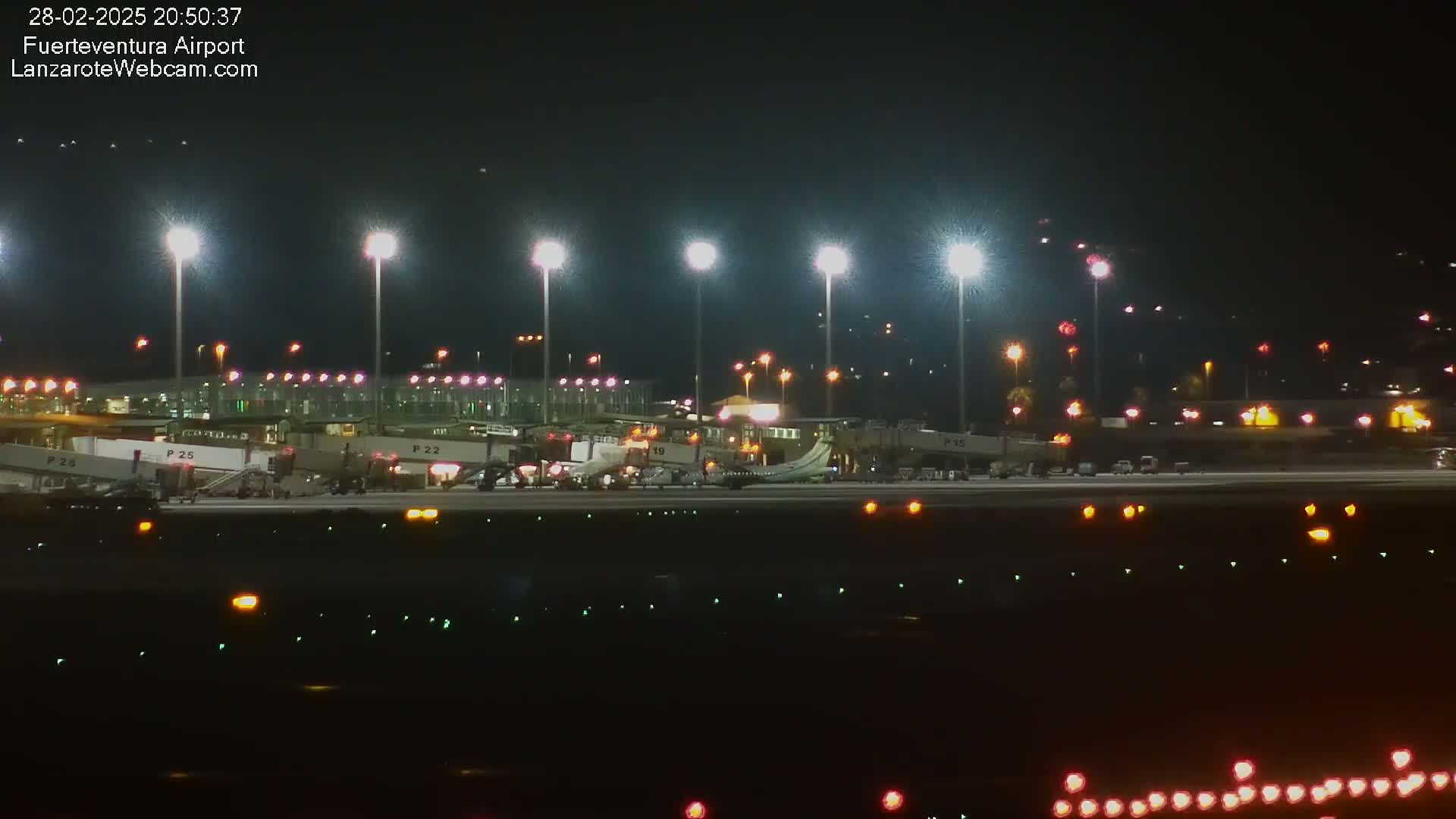 A brightly lit airport tarmac at night shows several airplanes parked at gates with jet bridges attached.