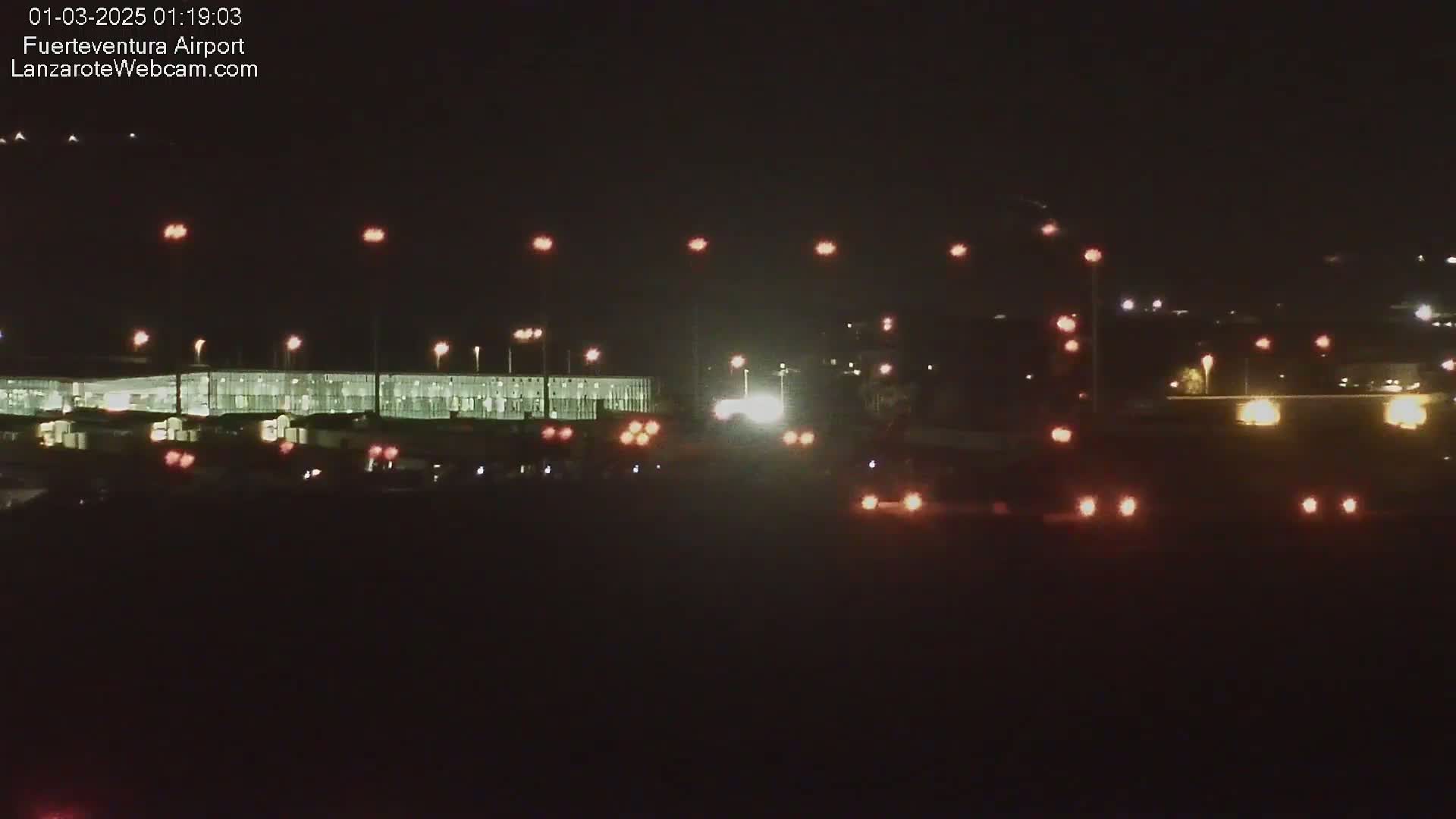 At night, a brightly lit airport terminal is visible with various vehicles and lights scattered in the foreground and background.