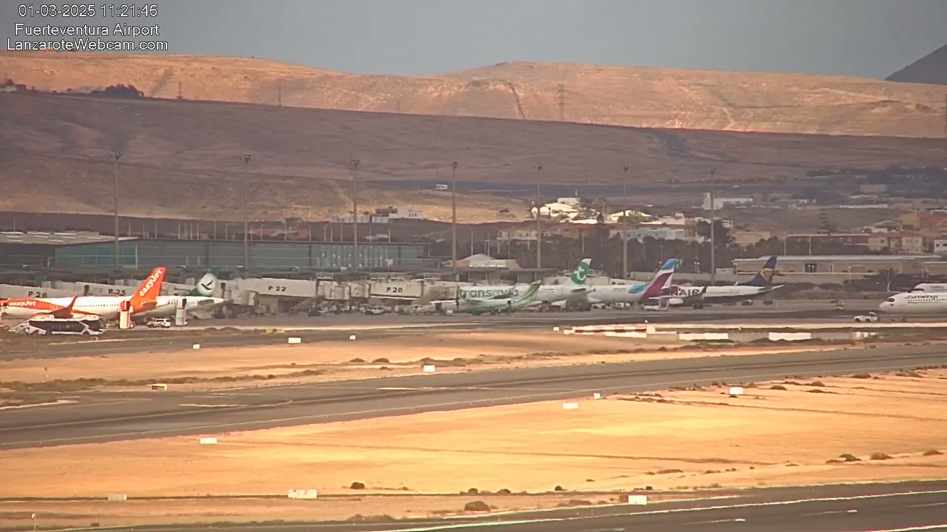 Several airplanes are parked at gates at an airport with a tan-colored landscape and hills in the background under a partly cloudy sky.