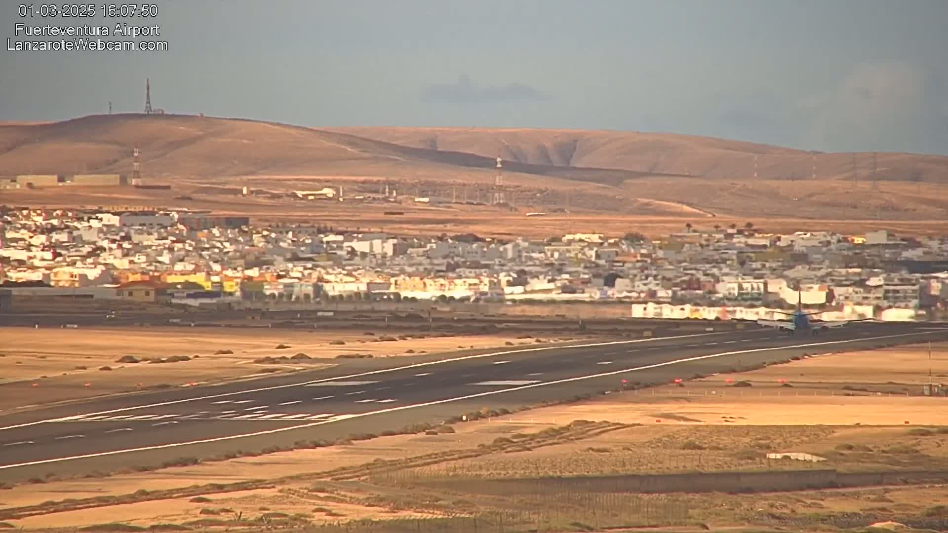 A plane is landing on a runway, with a town and arid hills visible in the background under a mostly clear sky.