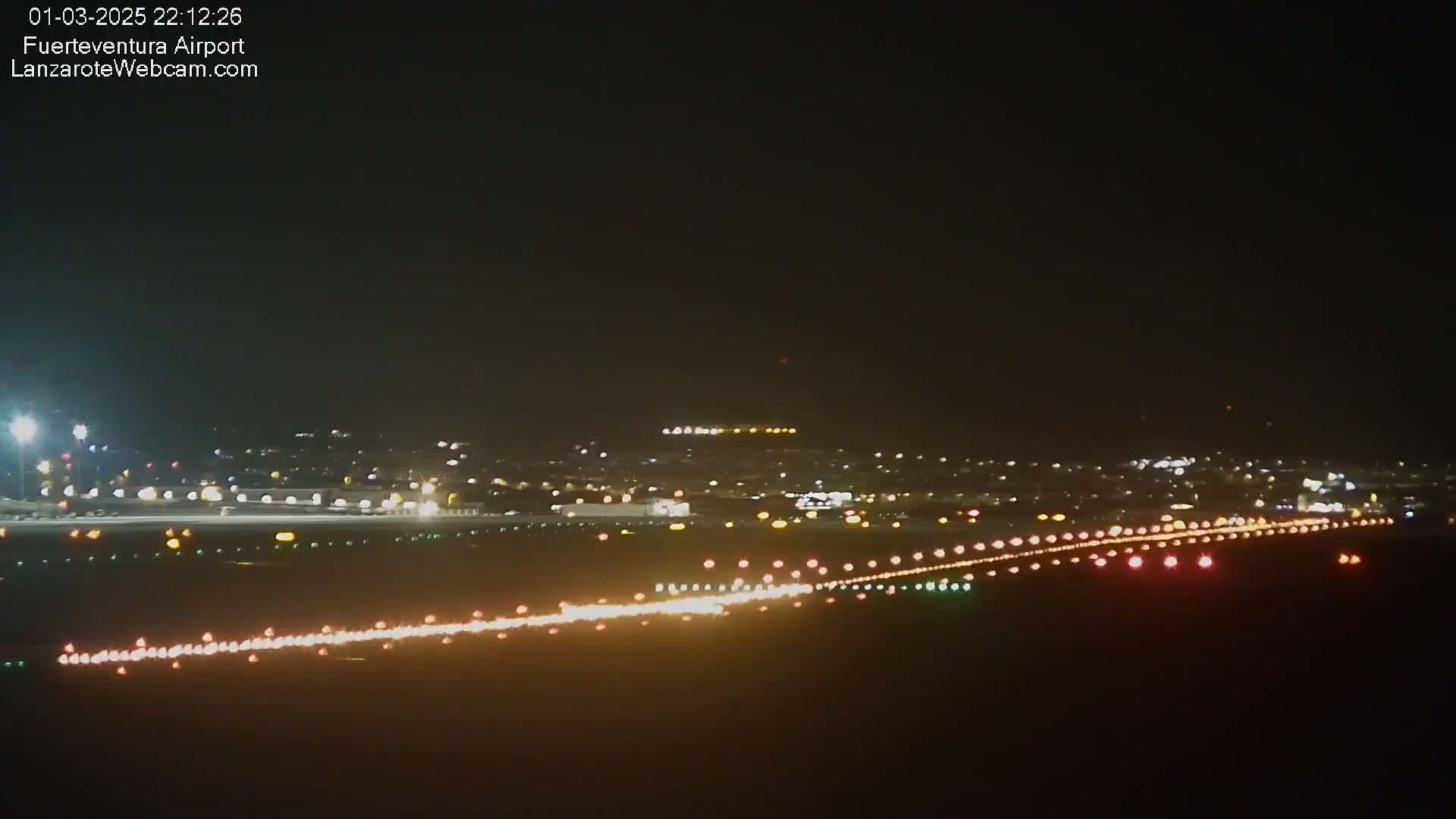 A nighttime long shot of an airport runway illuminated with lights, showing a city skyline in the background under a clear, dark sky.
