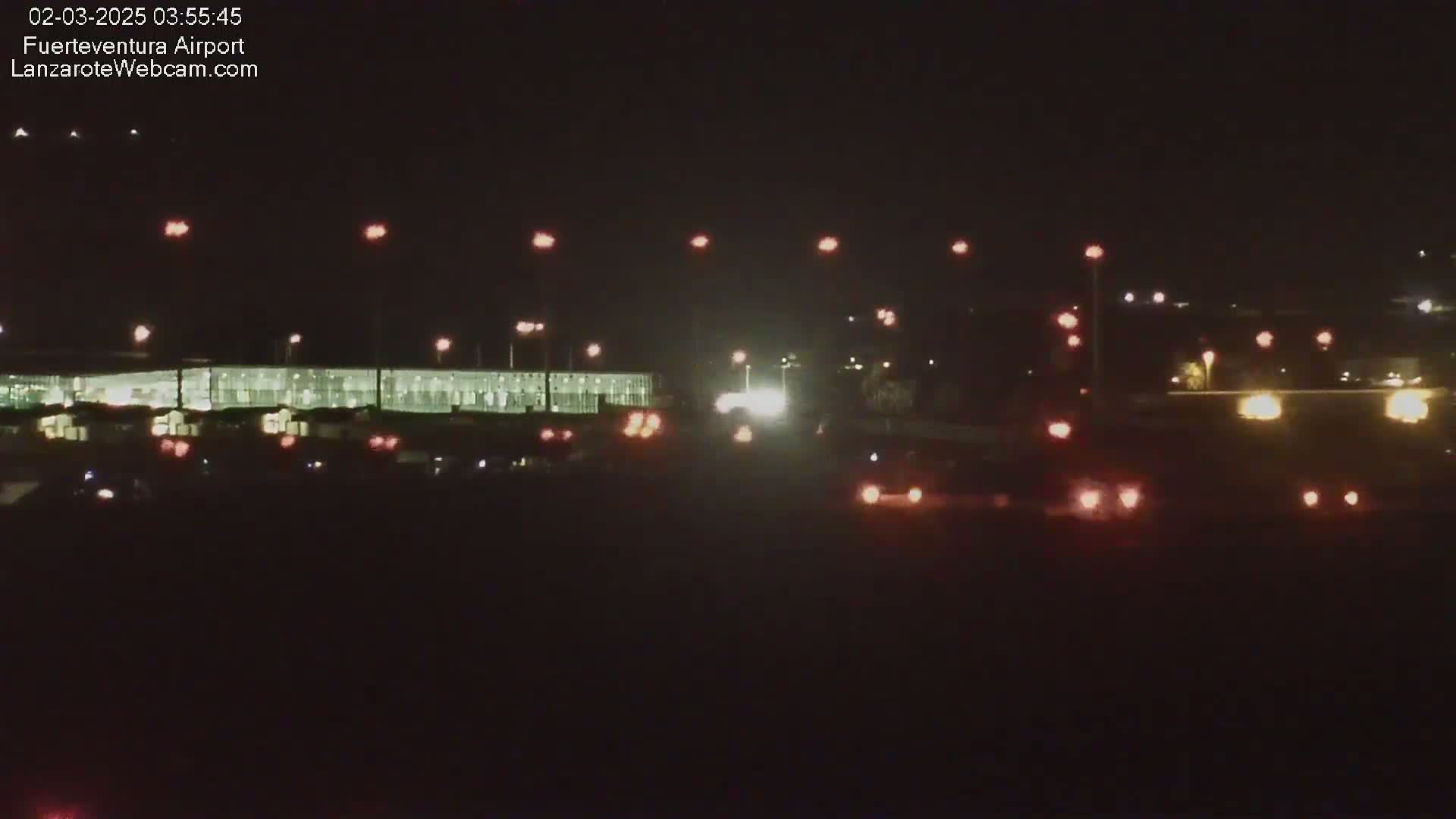At night, a long, brightly lit building is visible, with numerous lights scattered across a dark expanse in front of it.
