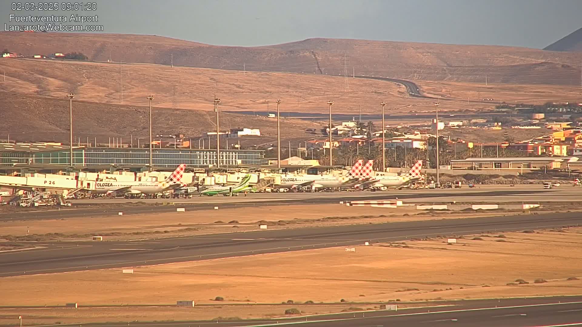 Several airplanes are parked at a gate at an airport under a hazy sky, with arid hills visible in the background.