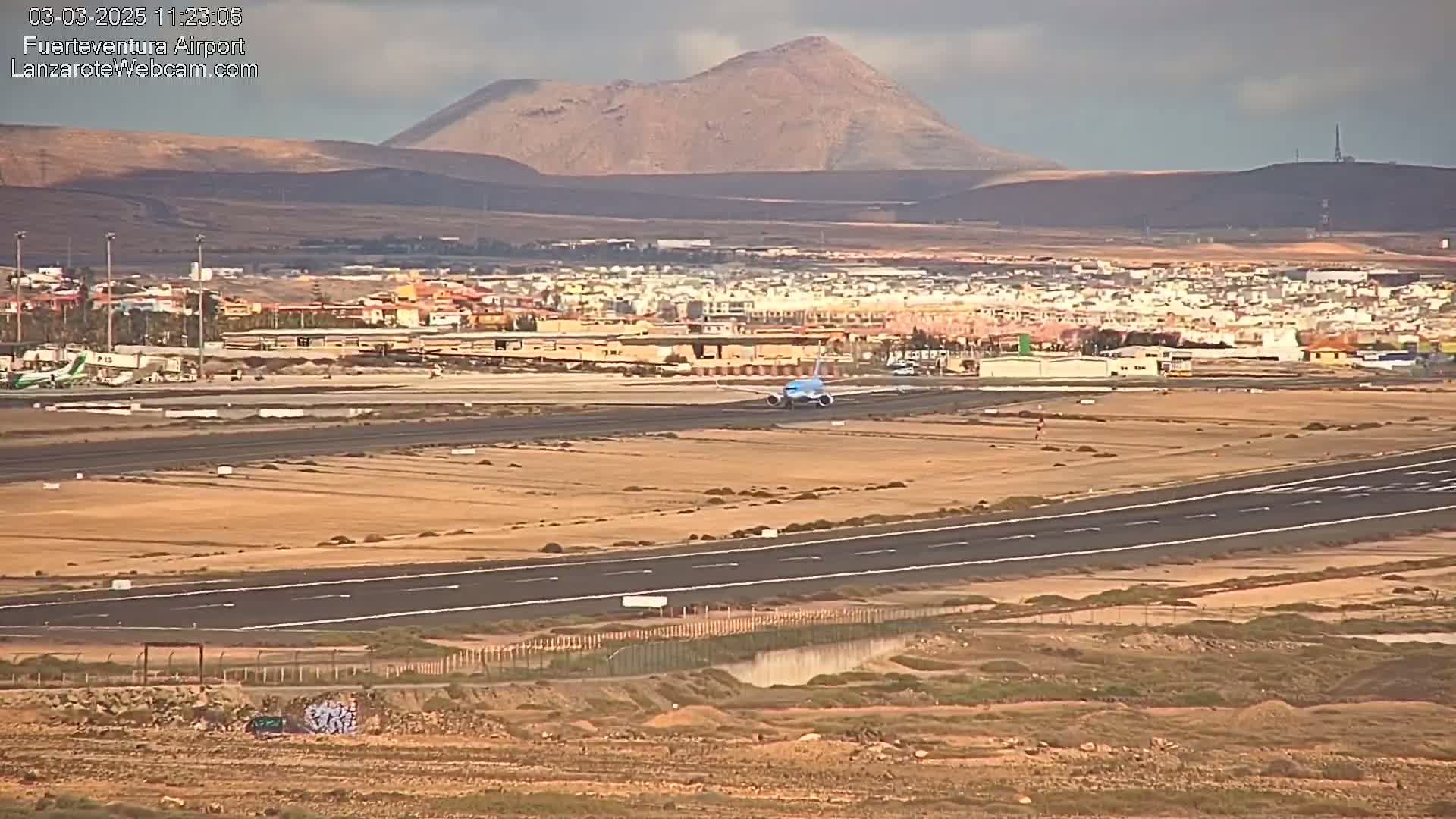 A light blue airplane taxis on a runway at an airport near a town and a large mountain under a mostly cloudy sky.
