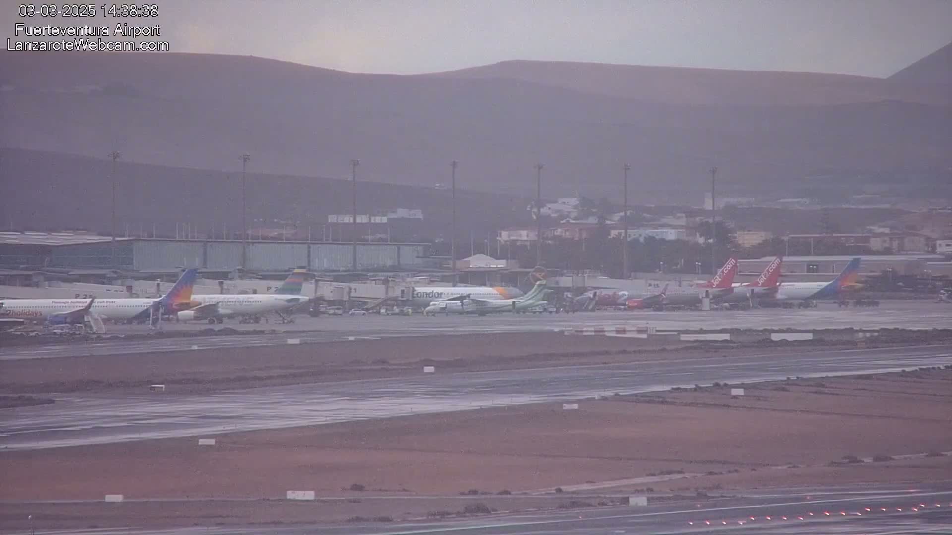 Several airplanes are parked at an airport on a hazy, overcast day with a mountain range in the background.