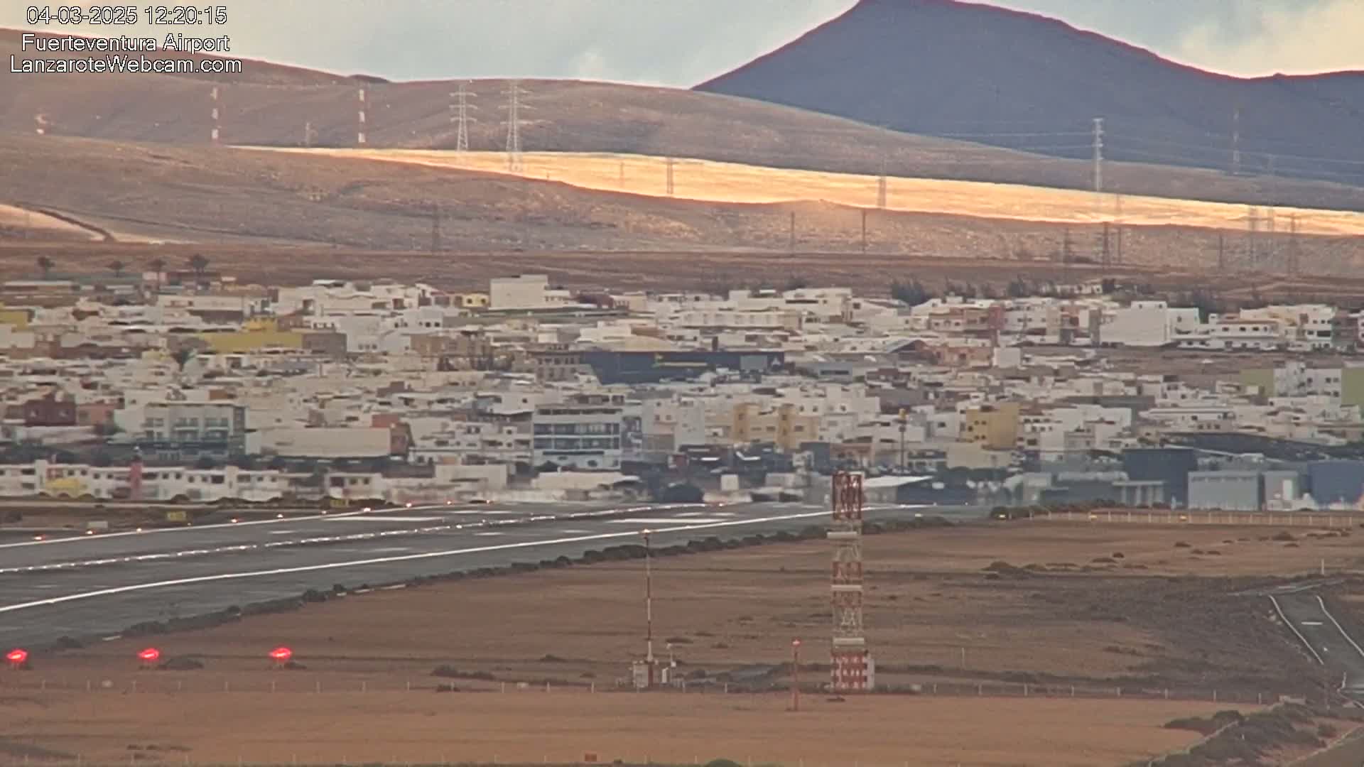 An airport runway with a town and a volcanic hill in the background under partly sunny conditions.