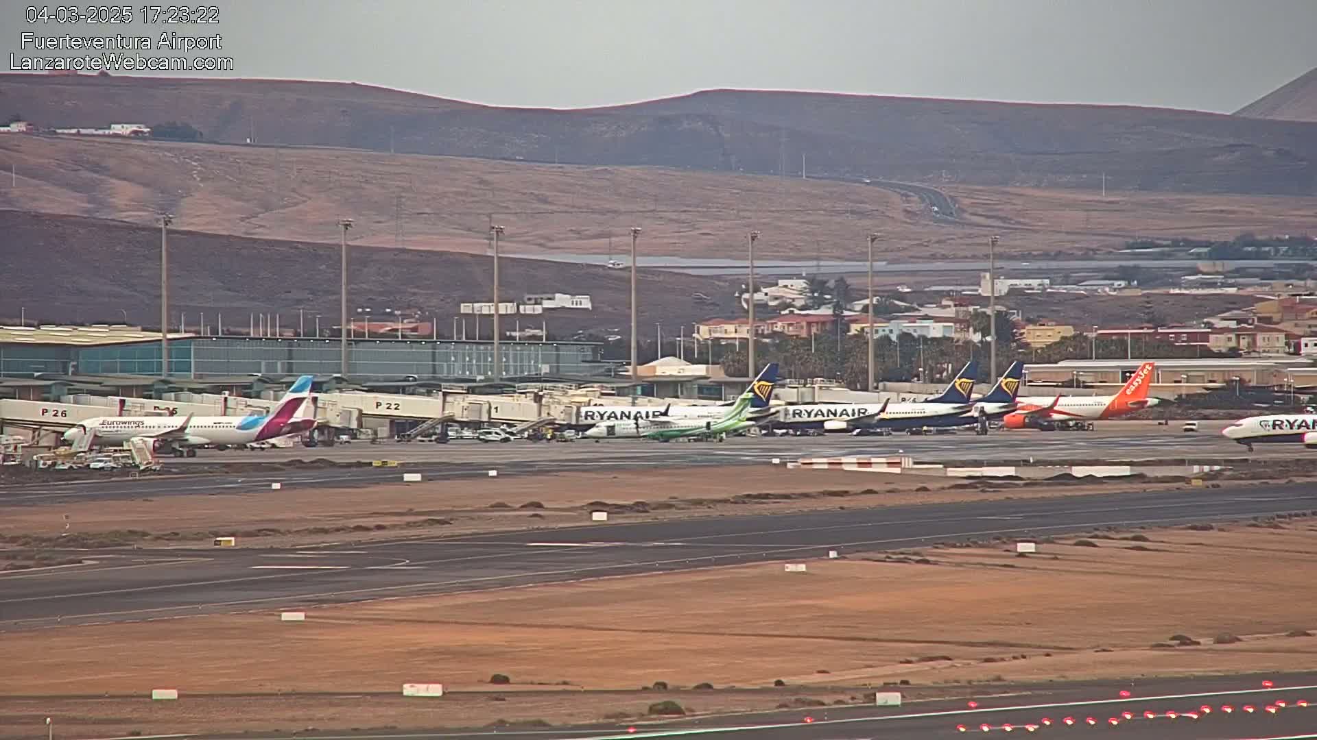 Several airplanes are parked at an airport gate under an overcast sky, with a hilly landscape visible in the background.