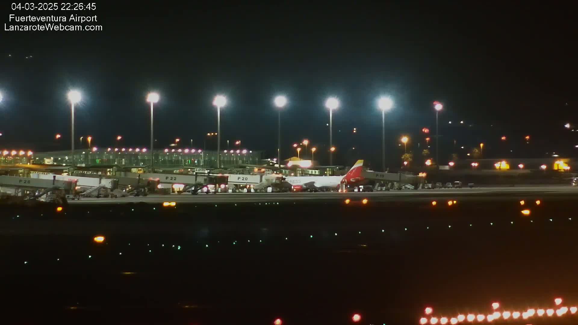 A large airplane is parked at a gate at night, illuminated by bright lights and surrounded by airport infrastructure.