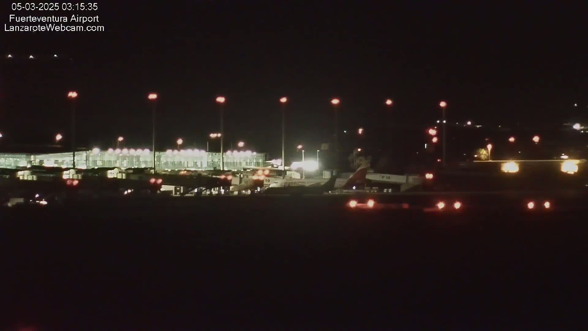 A nighttime view of an airport tarmac with airplanes parked at gates and ground support vehicles present under mostly dark skies.