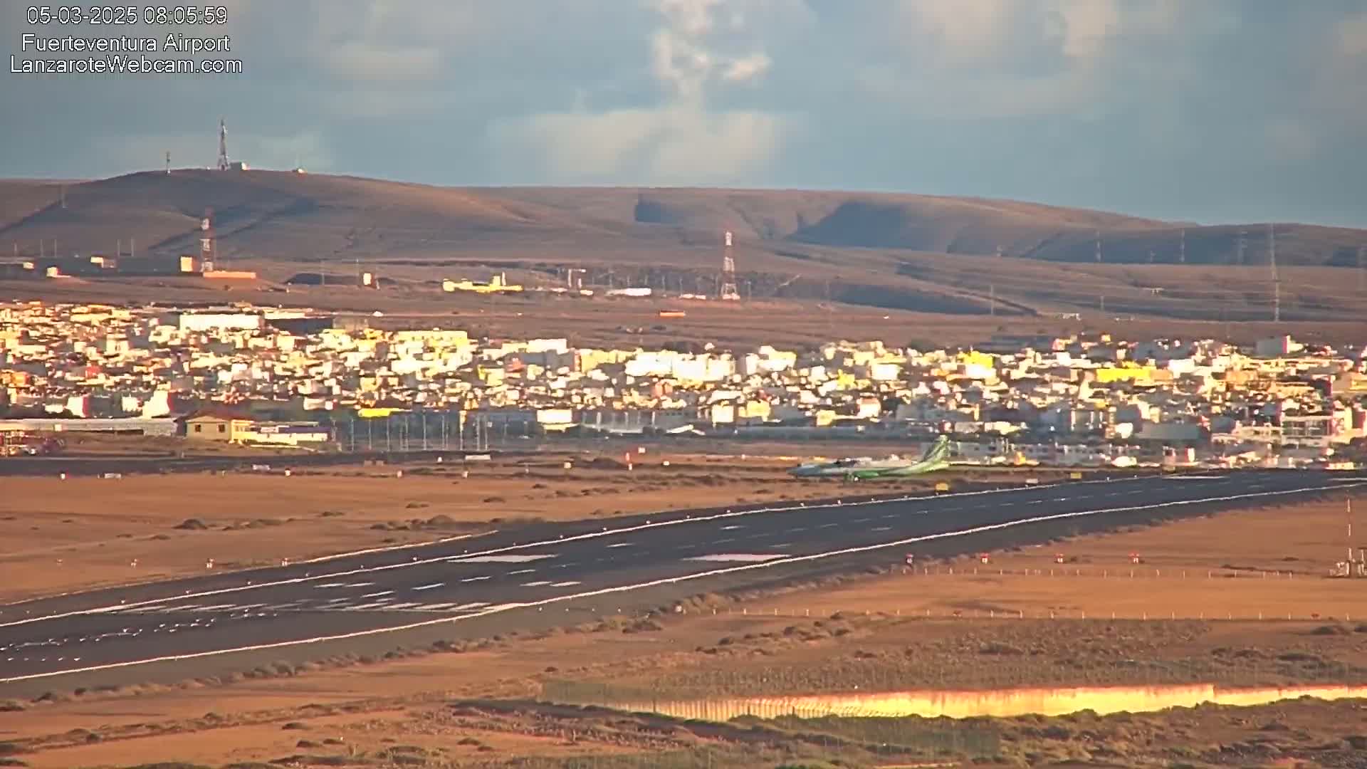 A plane taxis on a runway toward a town situated against a hilly, arid landscape under a partly cloudy sky.