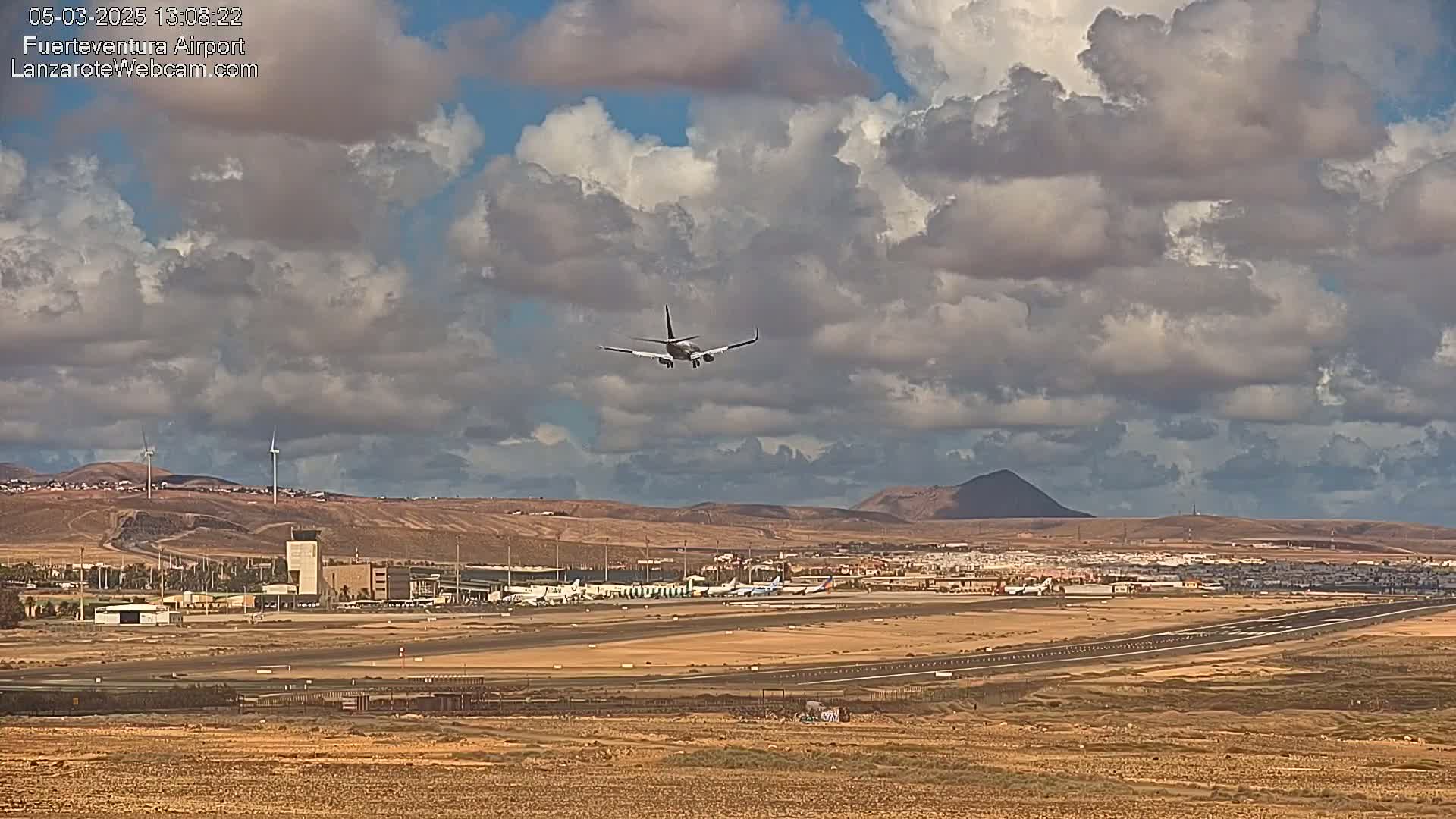 A commercial airplane is descending for landing over a flat, arid airport under a partly cloudy sky.