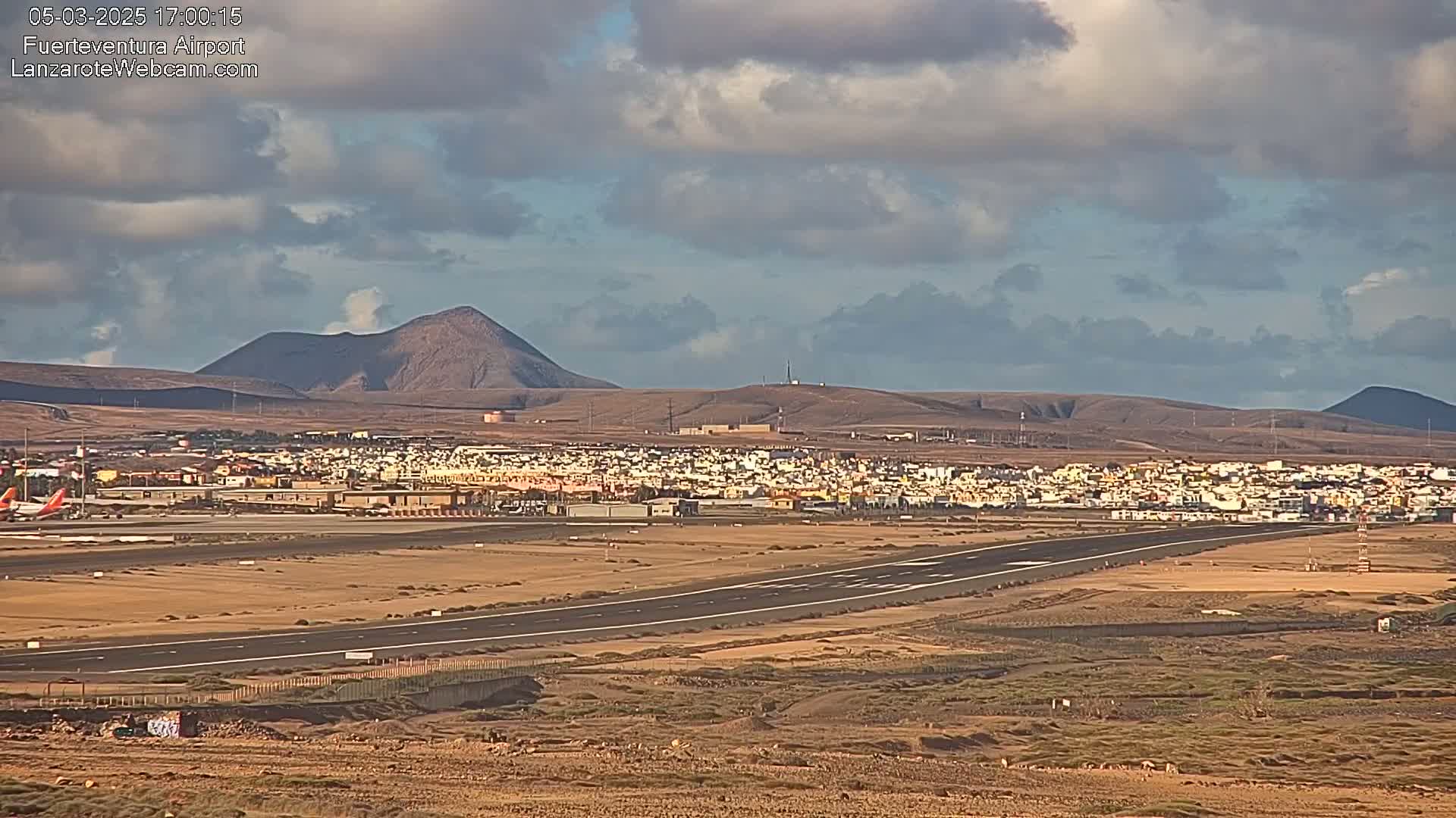 A runway at an airport is visible, with a town and volcanic hills in the background under a partly cloudy sky.