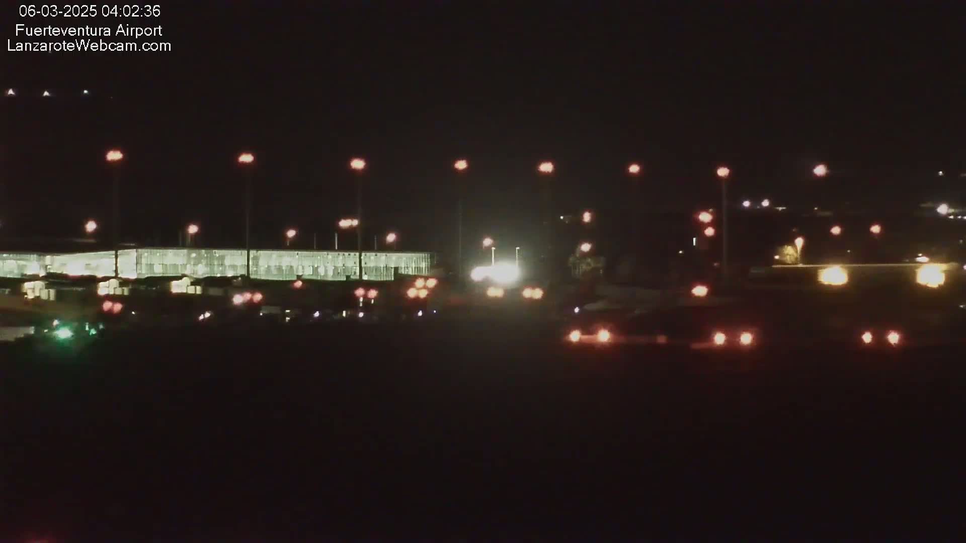 At night, a brightly lit airport terminal is visible, with numerous vehicles' headlights and taillights illuminating a roadway in front.