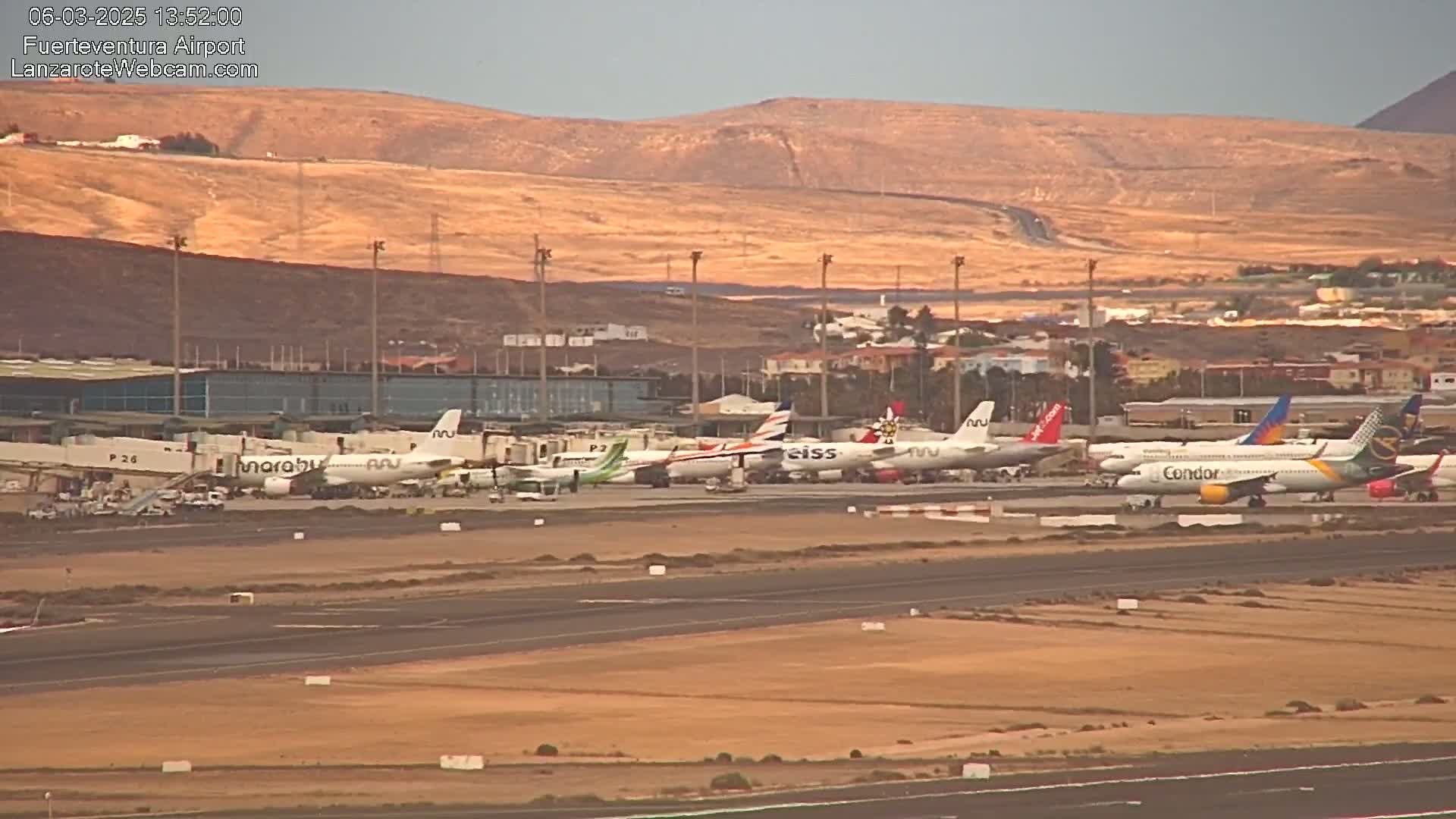 Several airplanes are parked at an airport apron under a hazy sky, with arid hills visible in the background.