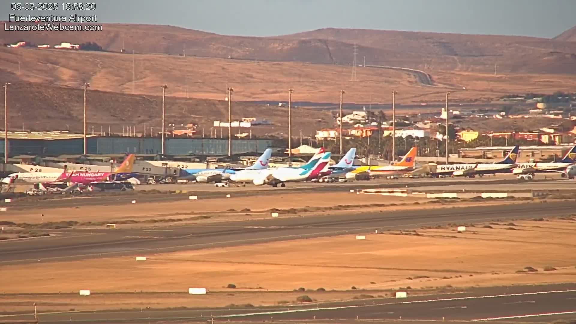 Multiple airplanes are parked on a tarmac at an airport under a clear, sunny sky with tan-colored hills in the background.