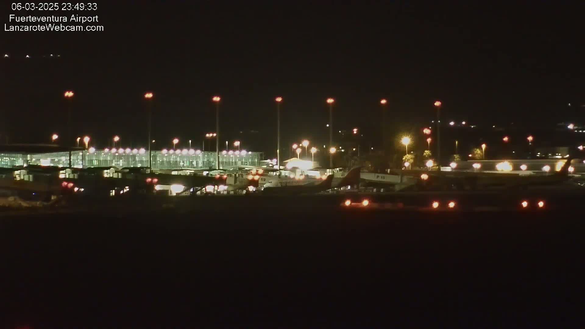 A nighttime view of an airport terminal and several airplanes on the tarmac, illuminated by numerous lights under a dark sky.