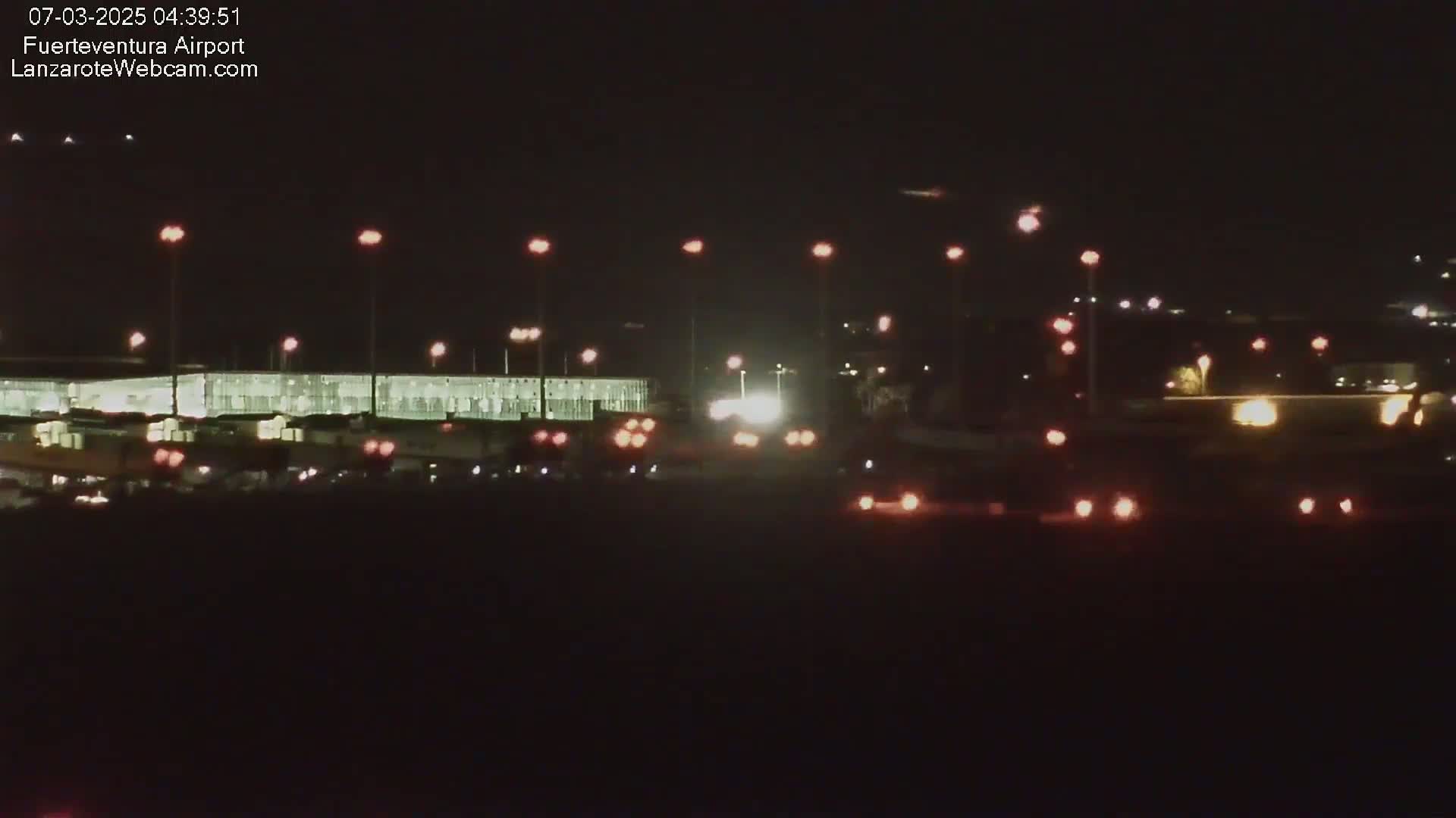 At night, a brightly lit airport terminal is visible, with numerous vehicles’ taillights present in the foreground.