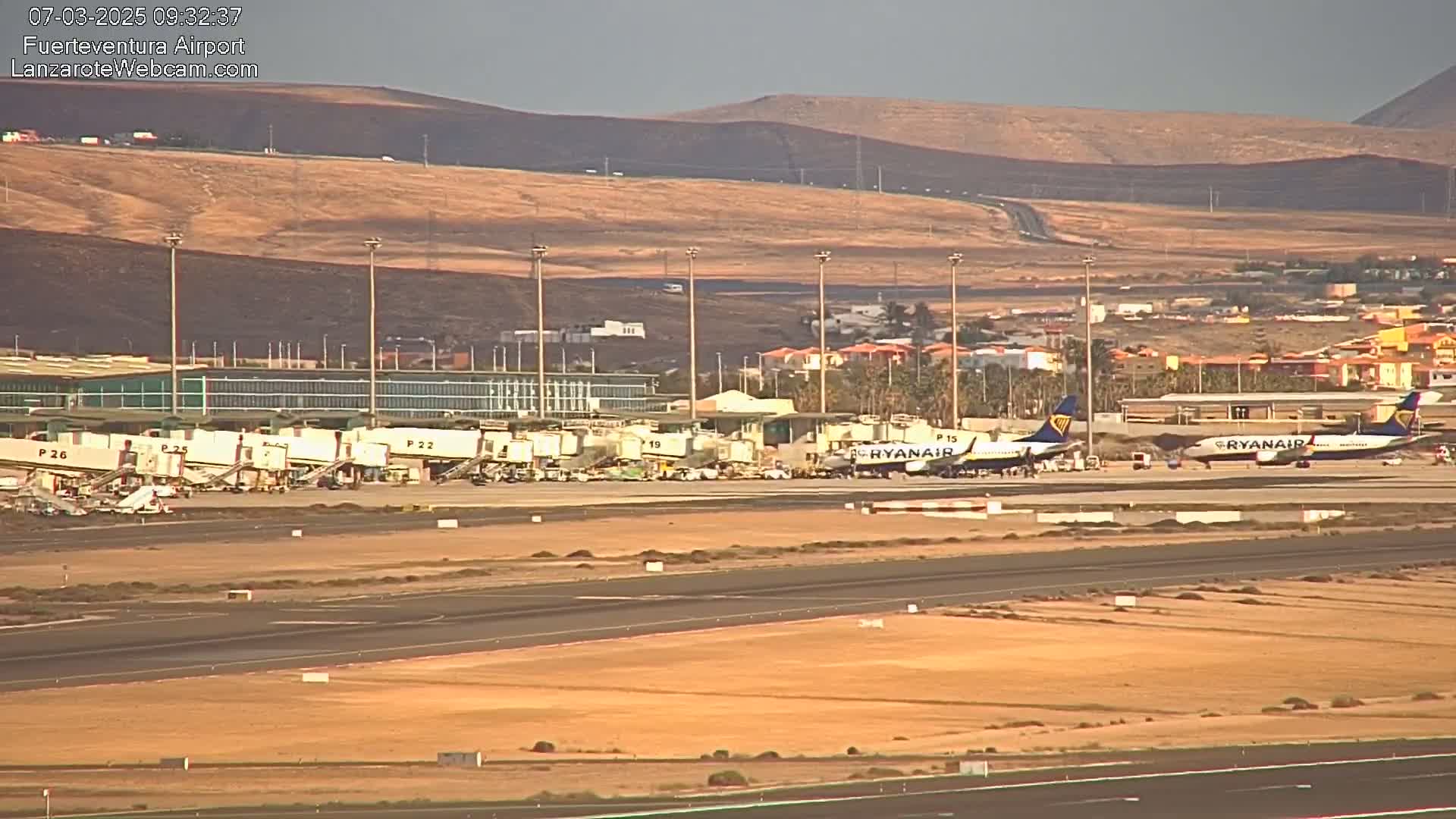Two Ryanair planes are parked at an airport tarmac under a mostly clear sky, with several jet bridges and airport buildings visible in the background, alongside a brown, hilly landscape.
