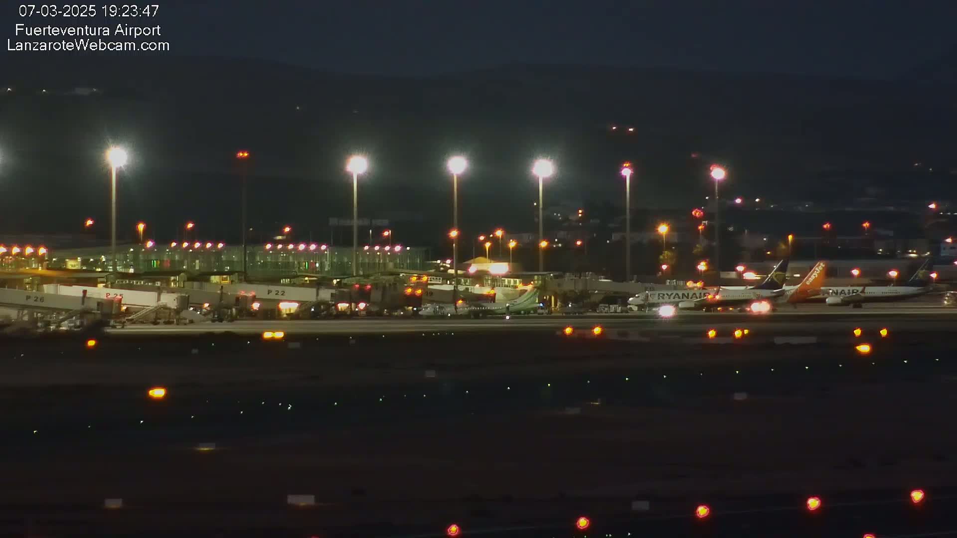 At night, a brightly lit airport tarmac shows several airplanes parked at gates, with ground support equipment visible.