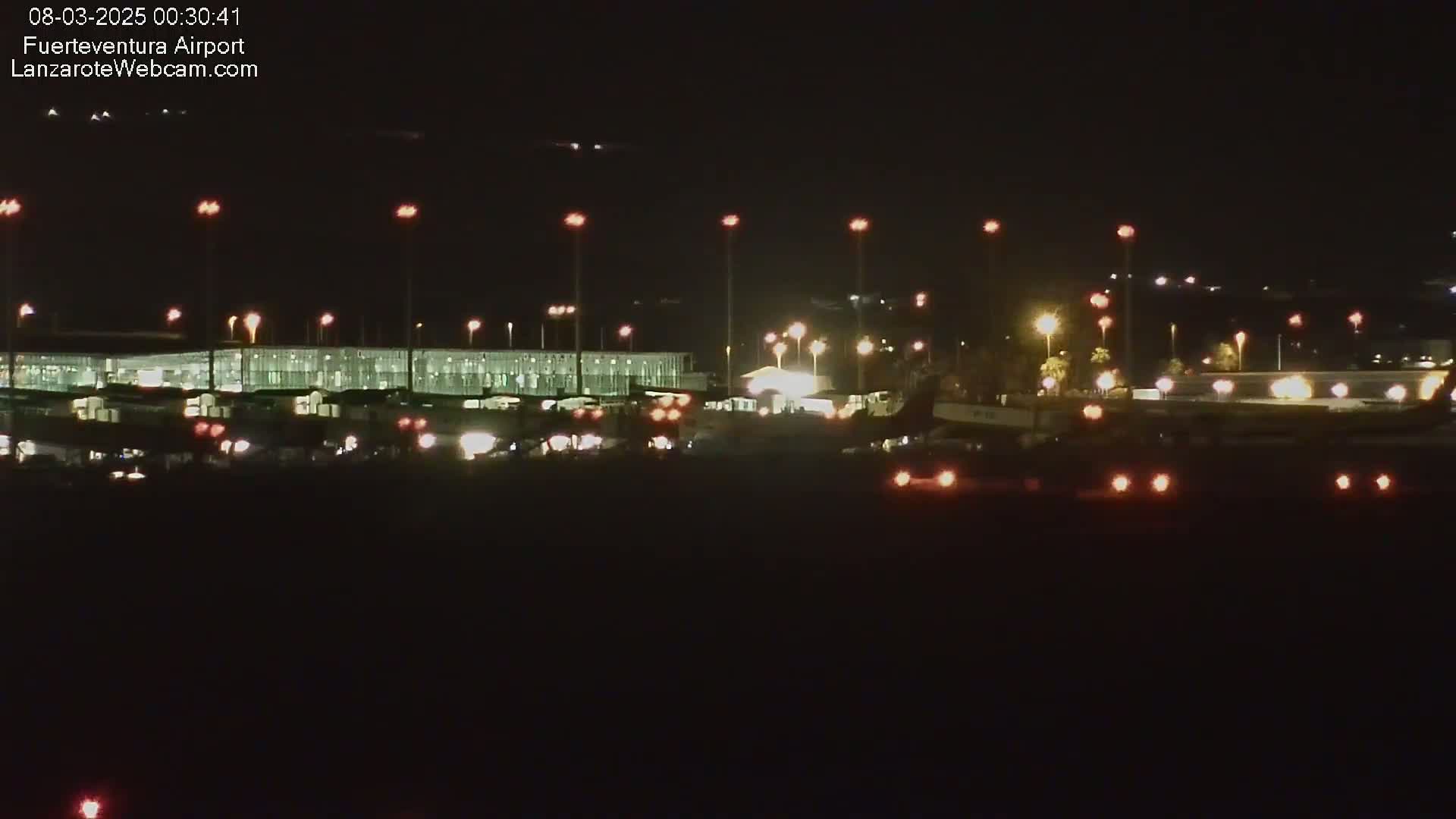 A brightly lit airport terminal and tarmac are visible at night, with several aircraft lights and runway lights illuminating the scene.