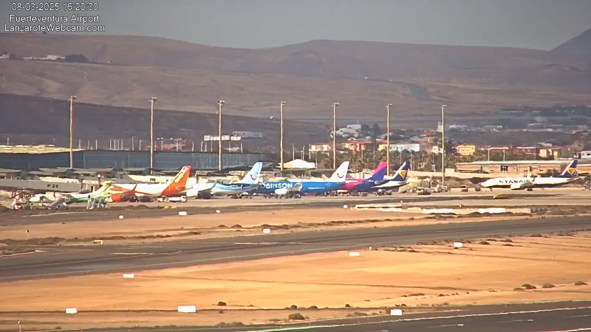 Several airplanes are parked on an airport tarmac under hazy, sunny conditions with a mountainous backdrop.