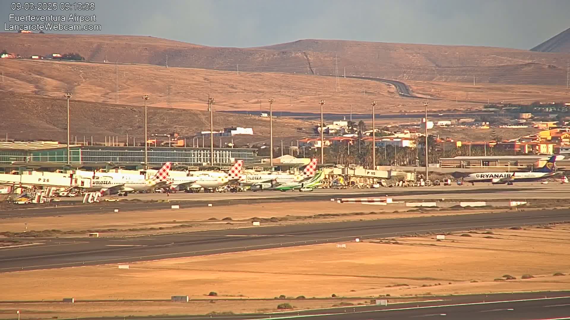 Several airplanes are parked at gates at an airport against a backdrop of dry, brown hills under a mostly clear sky.