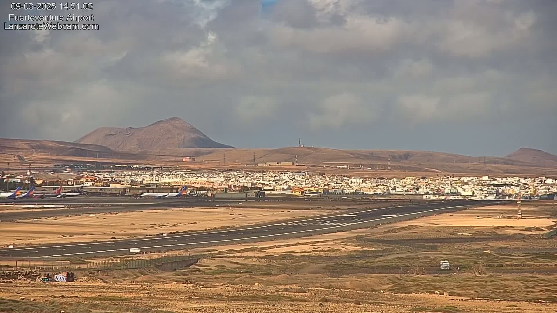 An airport runway and several airplanes are visible, with a town and volcanic hills in the background under a mostly cloudy sky.