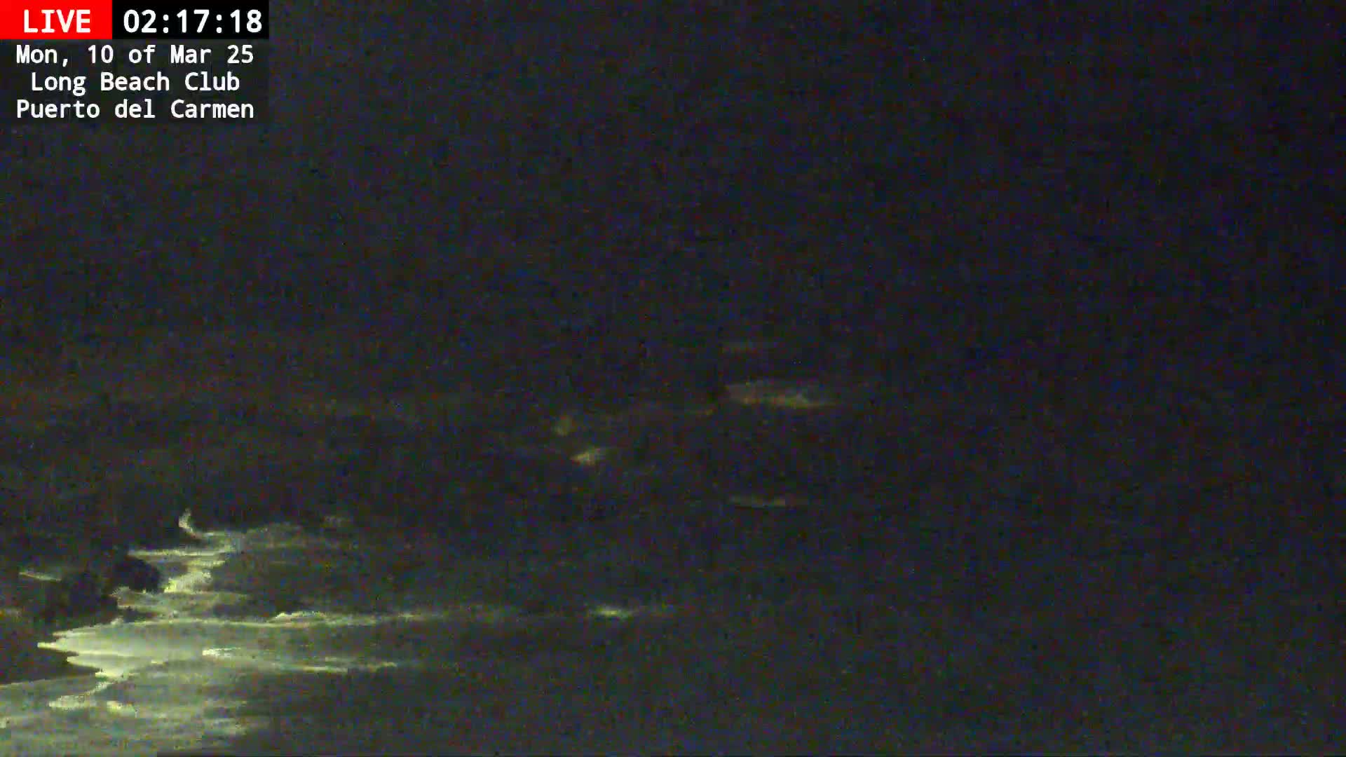 A dark, nighttime view of the ocean shows faintly glowing waves under a mostly cloudy sky.