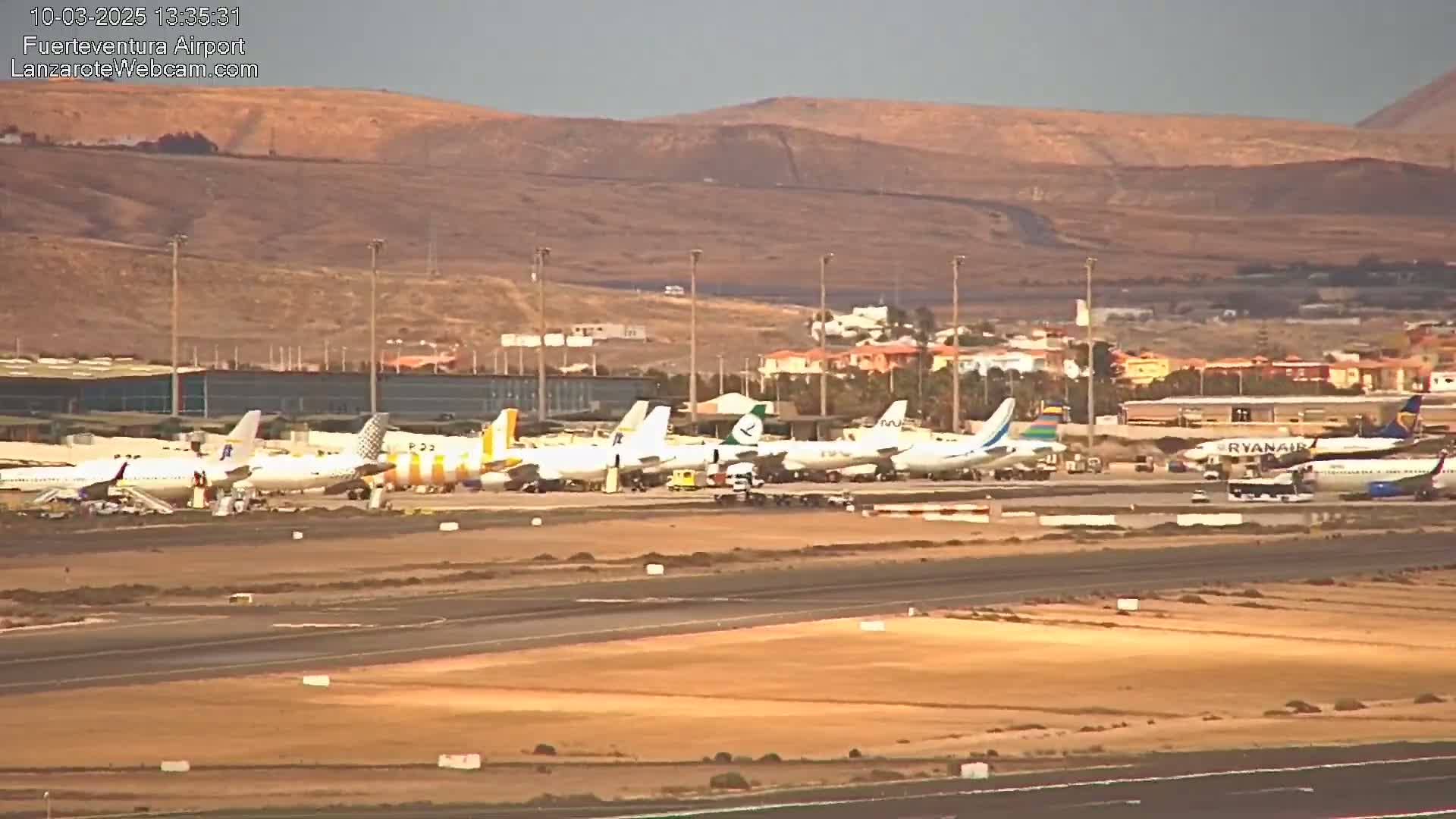 A long row of various passenger airplanes sits on an airport tarmac under a hazy sky, with desert-like hills visible in the background.