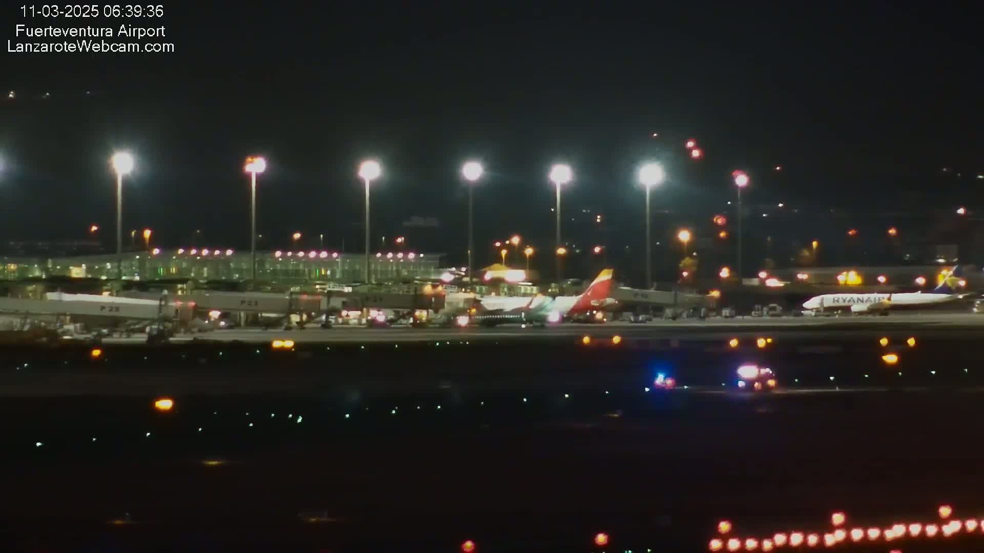 A nighttime view of an airport tarmac with several airplanes, including a Ryanair plane, near the gates, with bright lights illuminating the scene.