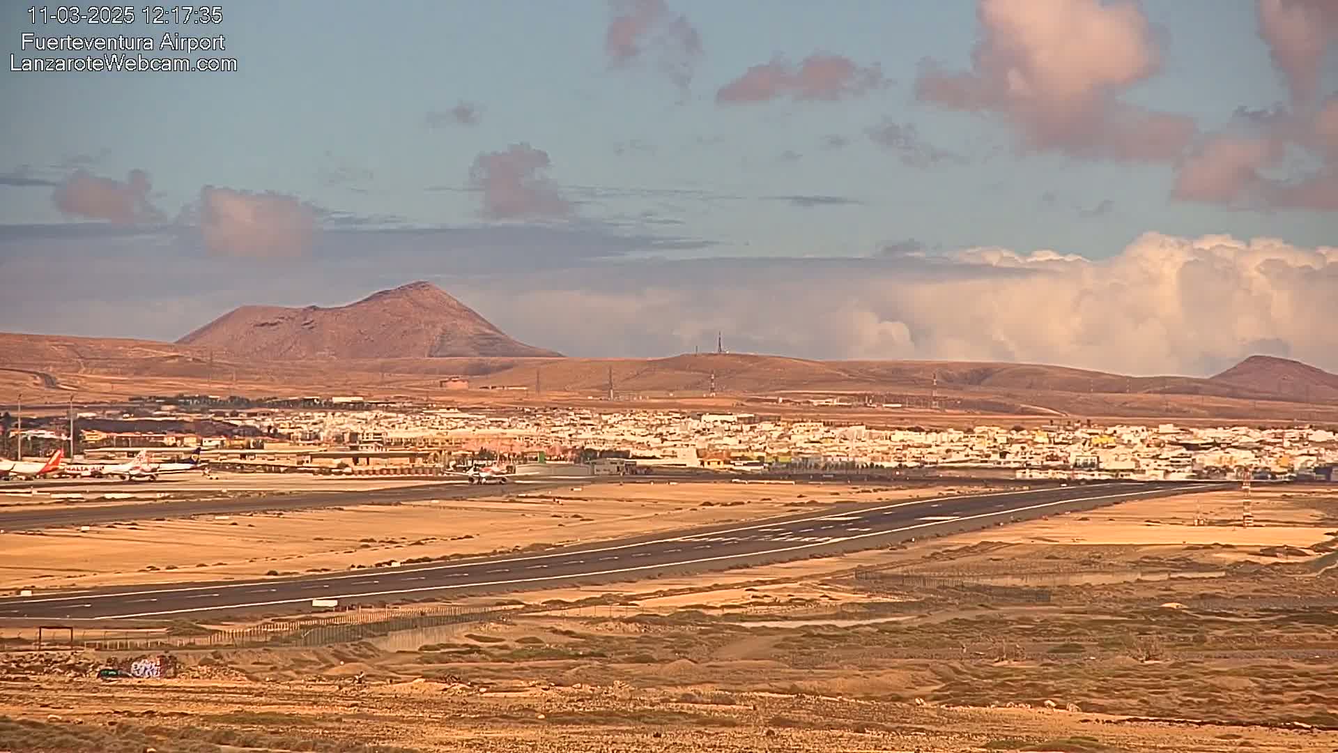 A long runway at an airport sits below a partly cloudy sky and in front of a town nestled between arid hills.