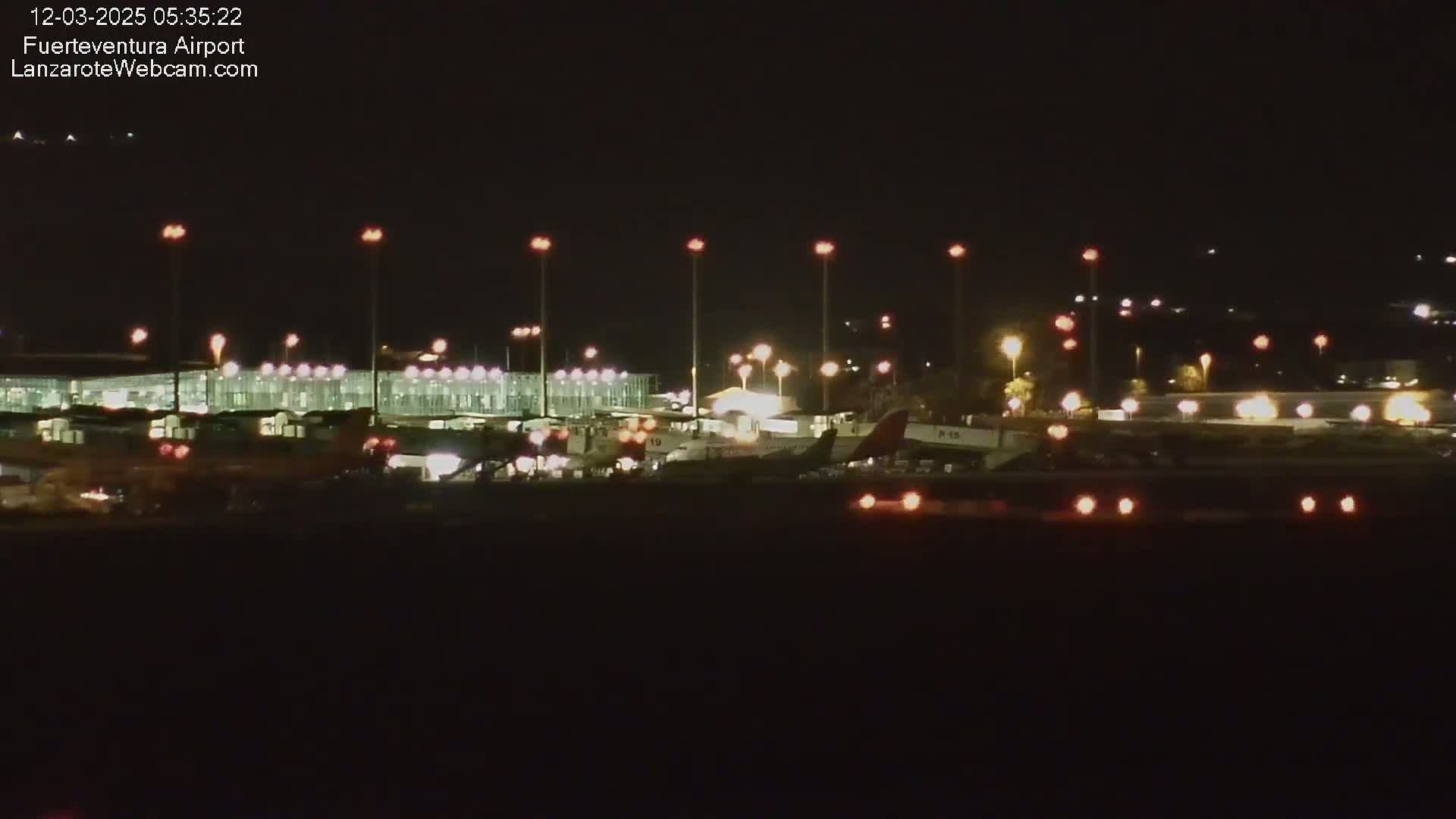 A nighttime view of an airport tarmac with several airplanes parked near a brightly lit terminal building under a dark sky.