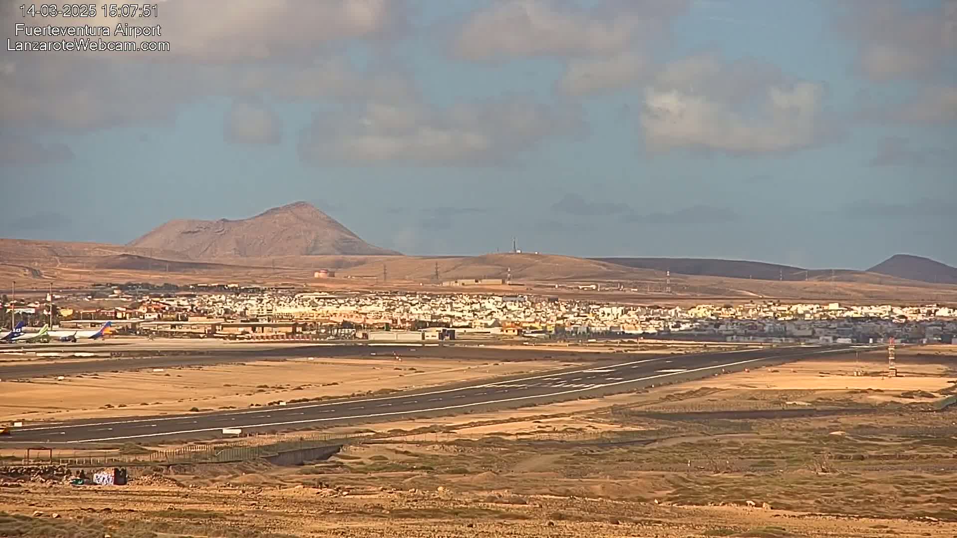 An airport runway stretches across a flat, arid landscape under a partly cloudy sky, with mountains visible in the distance.