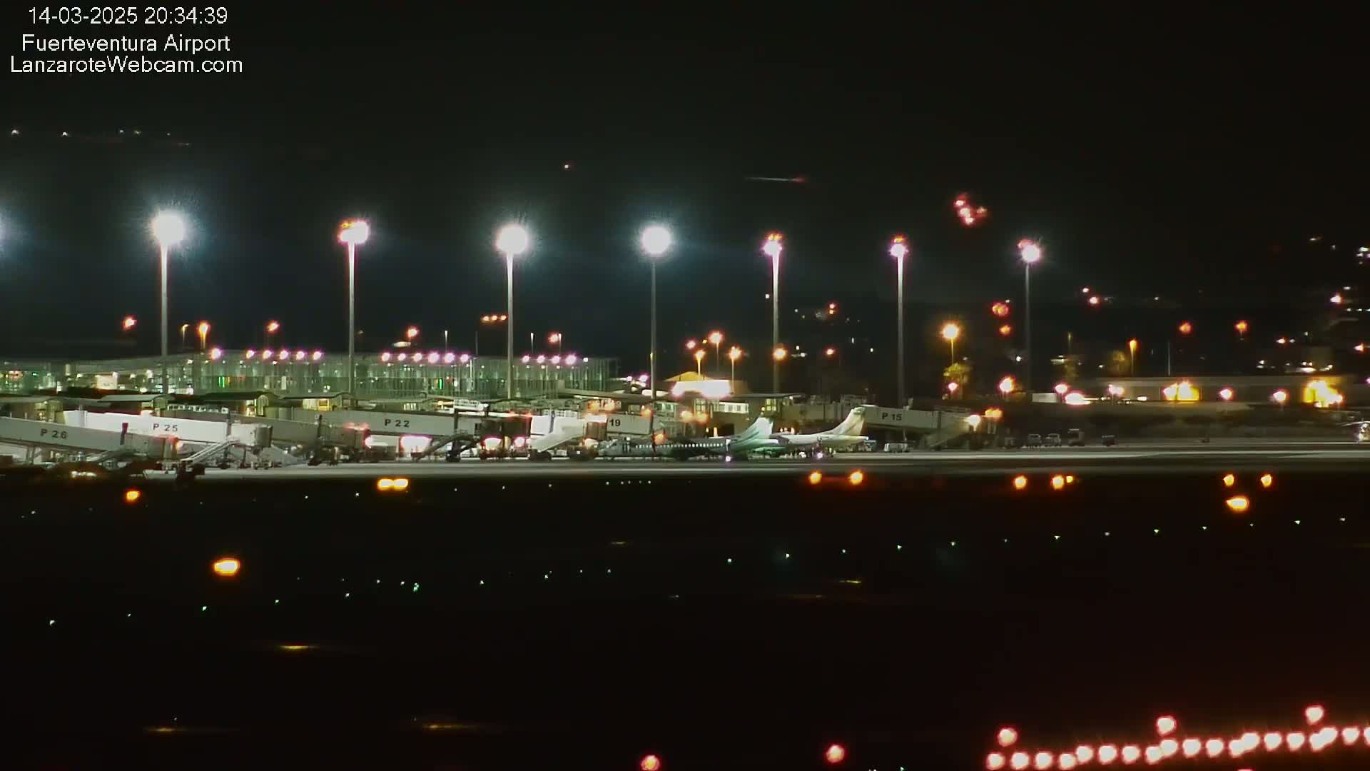 A brightly lit airplane is parked at a gate at night at an airport with several other planes visible in the distance.