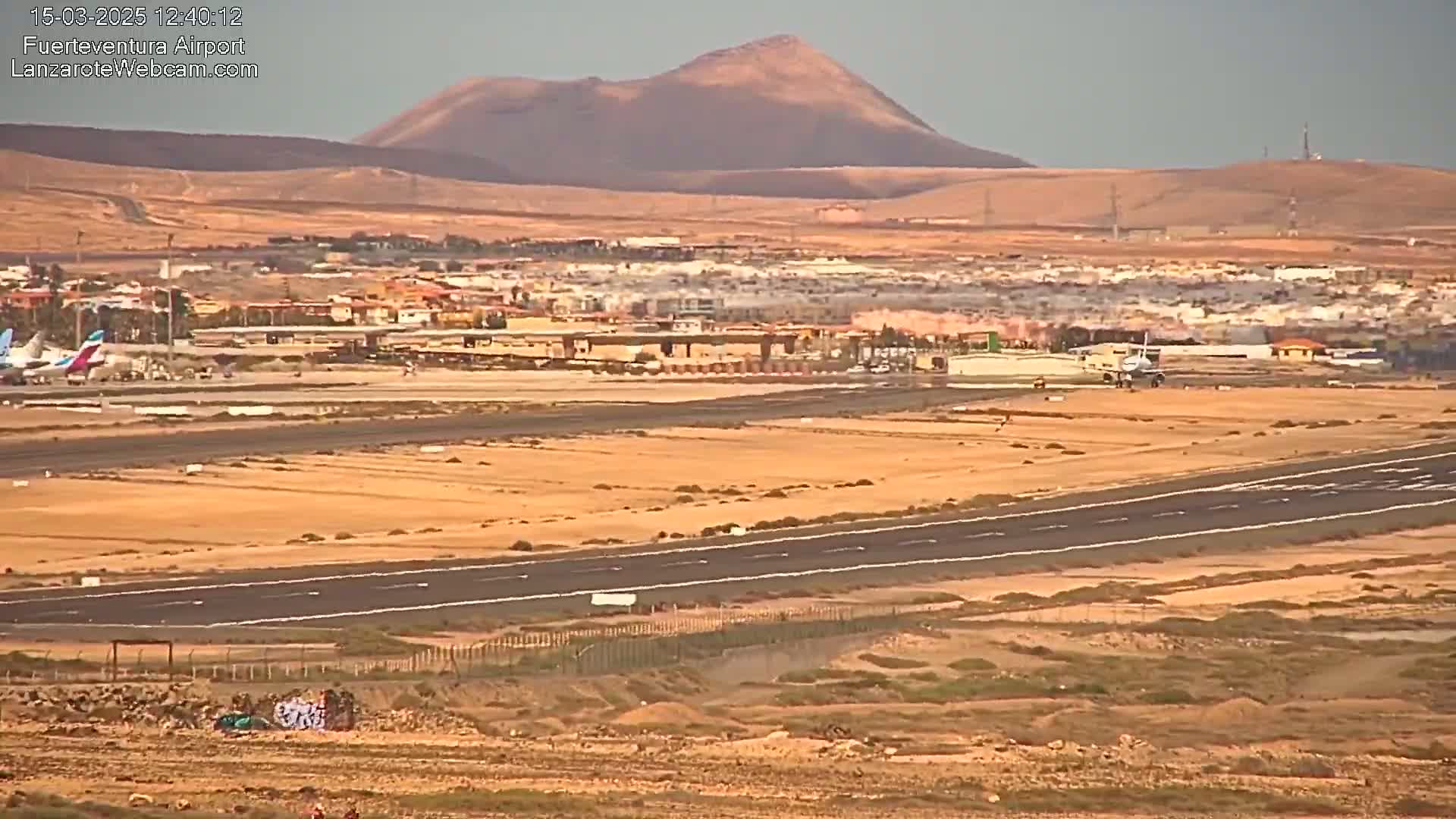 A long runway at an airport next to a town, with a hazy mountain in the background under a mostly clear sky.
