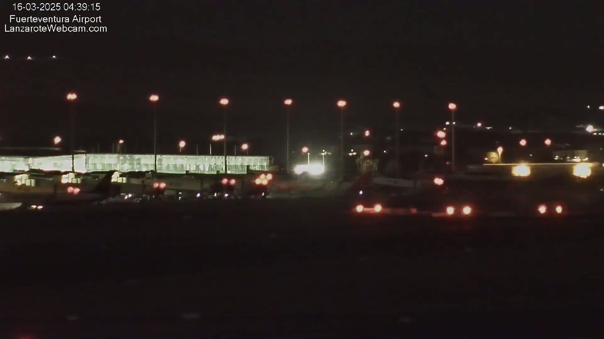 At night, an airport tarmac shows several vehicles and an airplane near a brightly lit terminal building under a dark sky.
