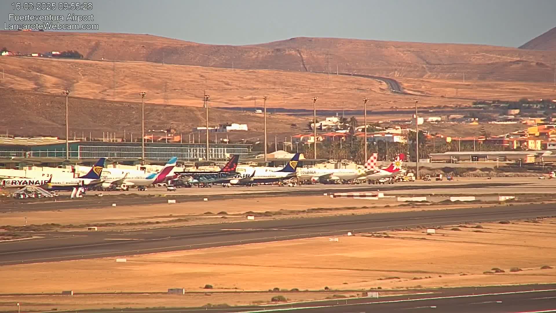 Several passenger airplanes are parked on an airport tarmac under a clear, sunny sky with arid, tan-colored hills in the background.